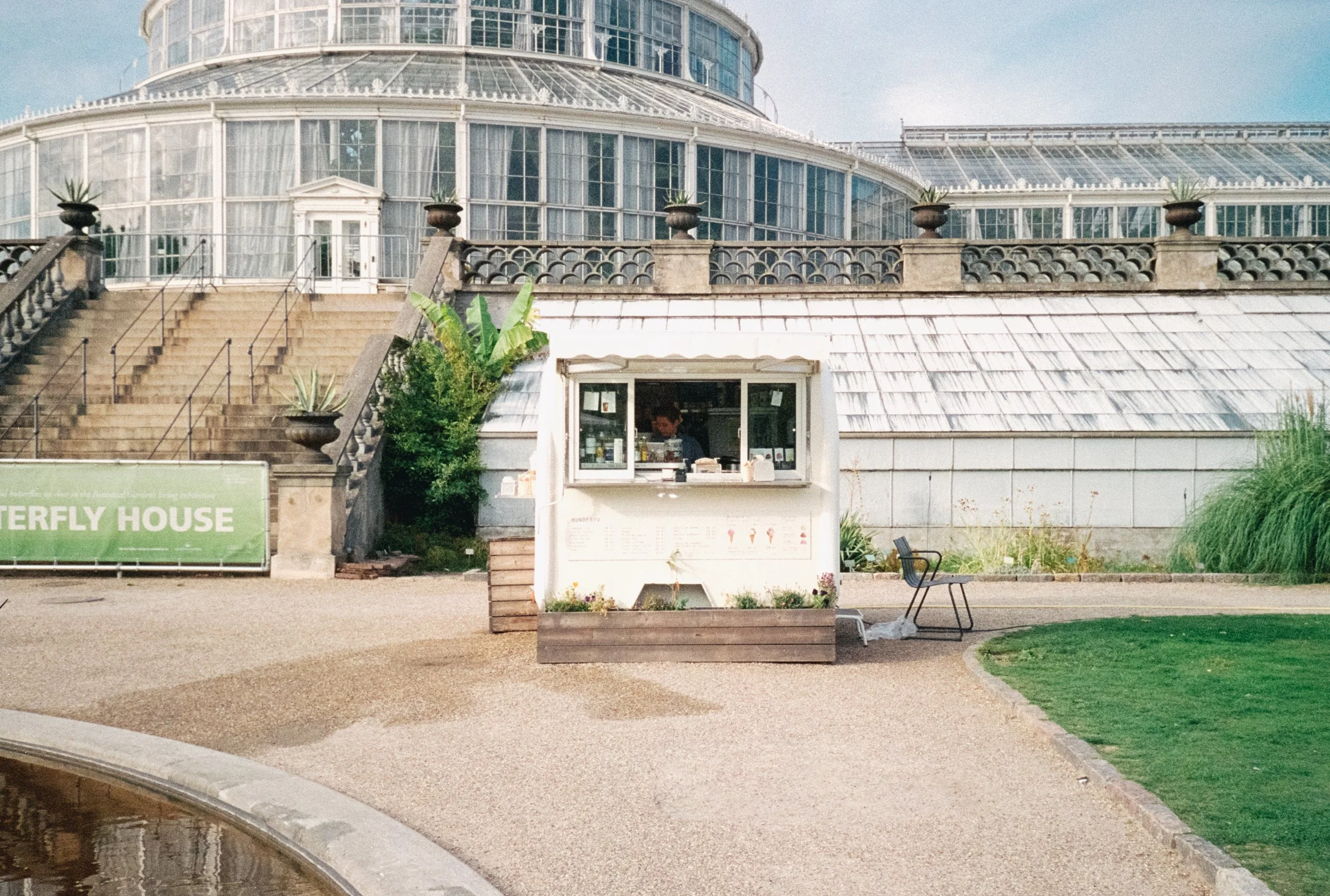 A small stand with a person inside selling ice cream cones in front of a glass conservatory or greenhouse with staircases and potted plants.