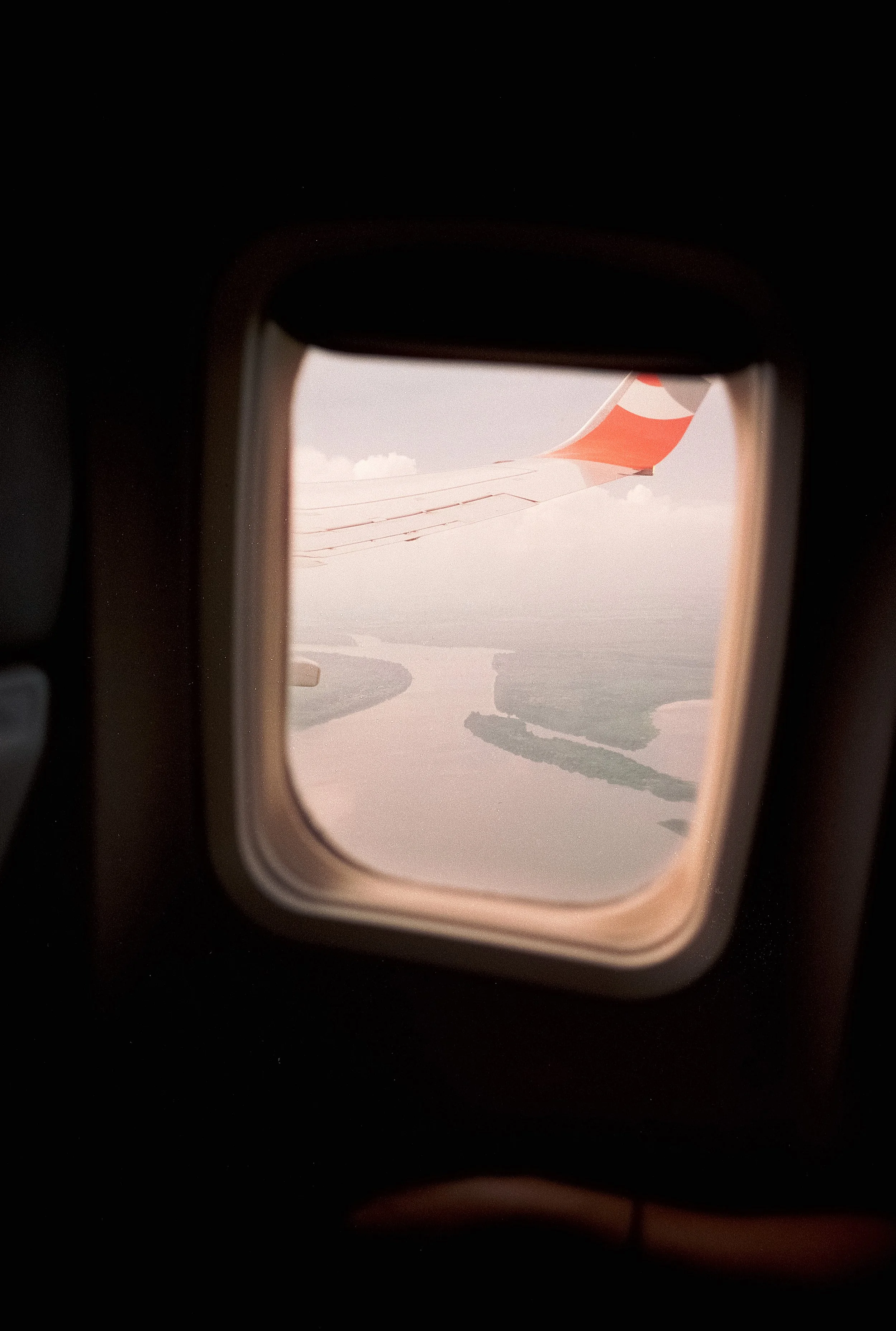 Photo taken from an airplane window showing part of the airplane wing and winglet with an airport runway and water bodies below on a cloudy day.