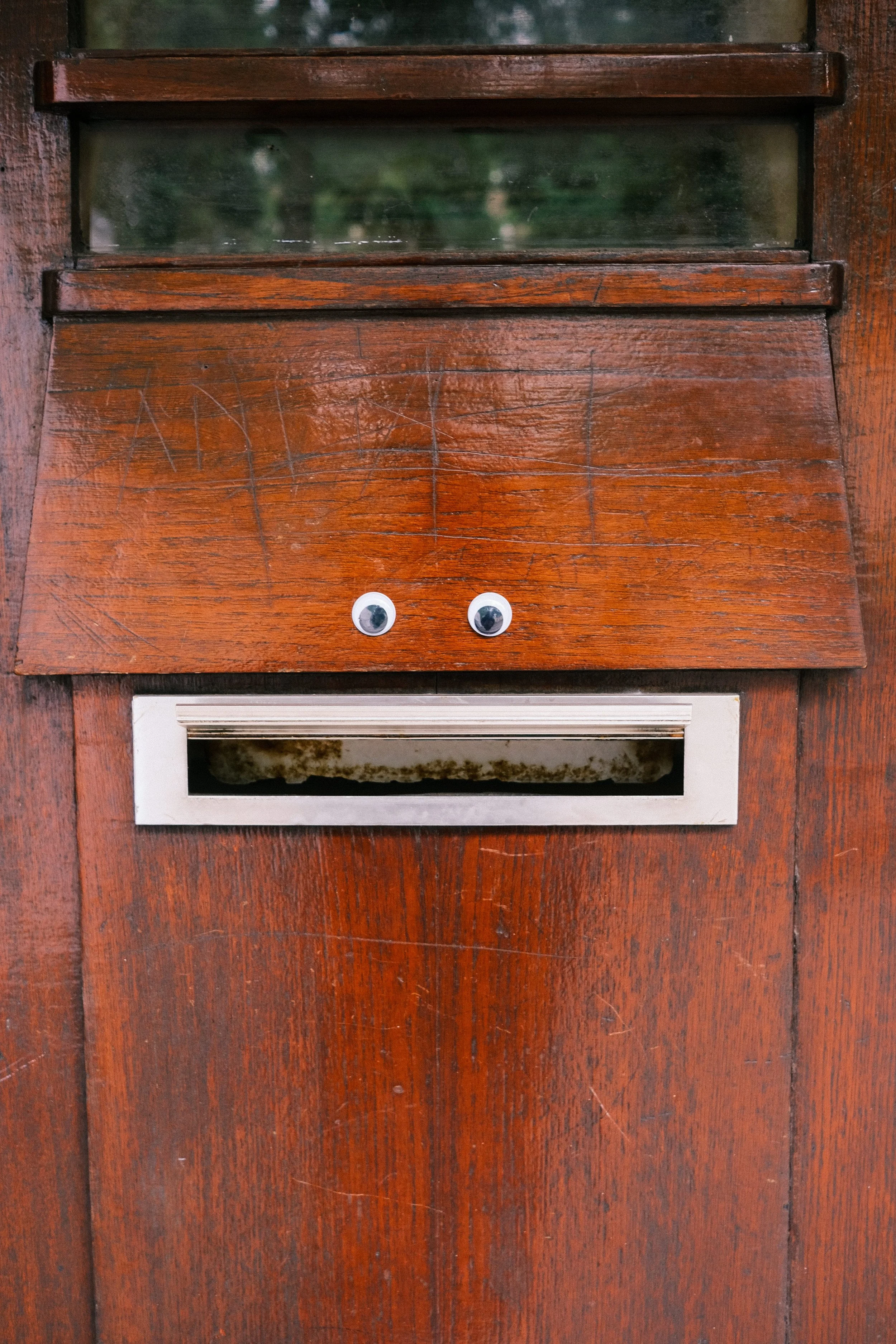 A wooden mailbox with googly eyes attached below the mail slot, mounted on a wooden surface.