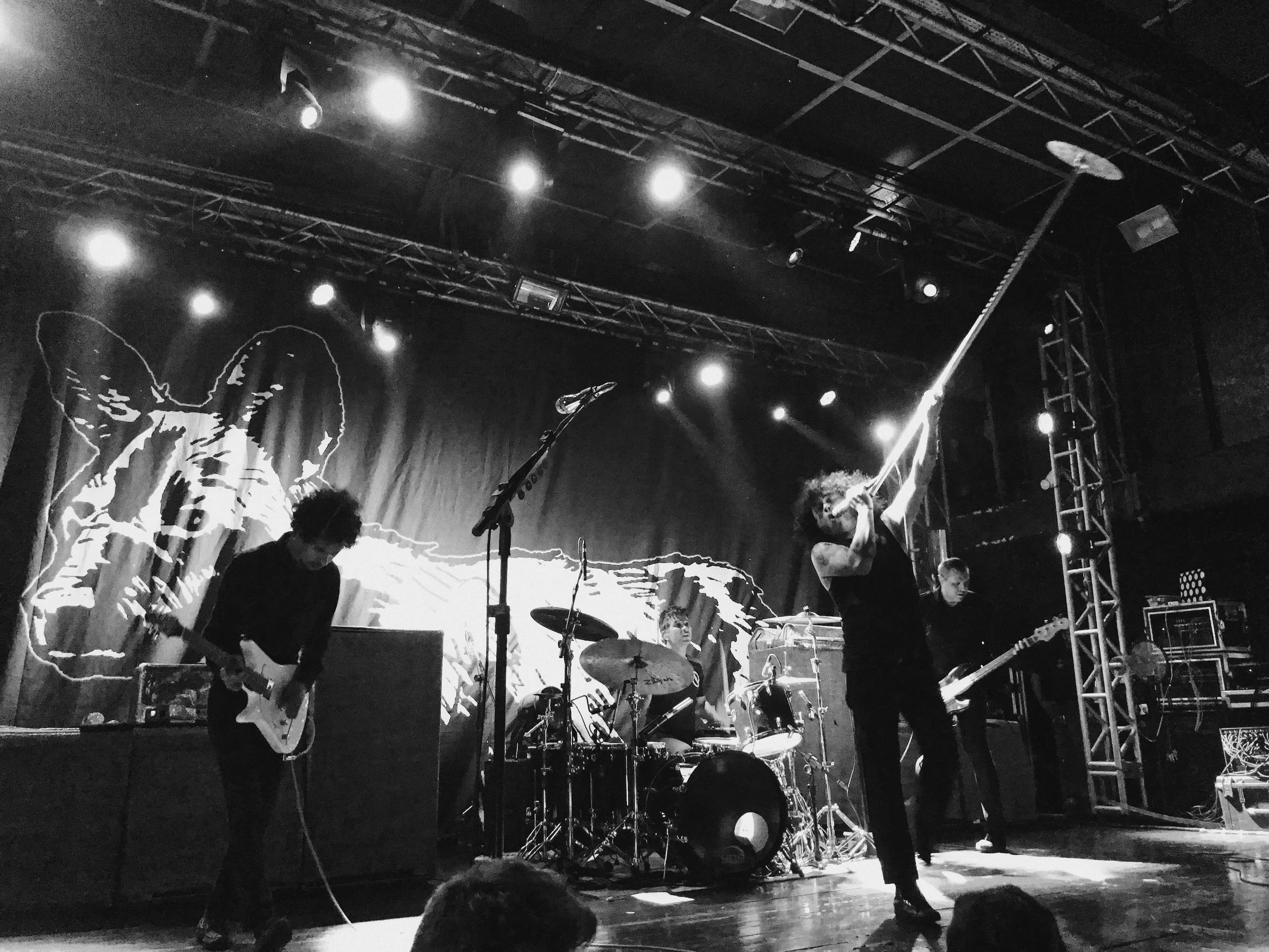 A black-and-white stage performance with a band. Musicians are playing guitars, drums, and keyboards. A singer is holding a microphone. Stage lighting illuminates the scene, and a large butterfly graphic is visible in the background.