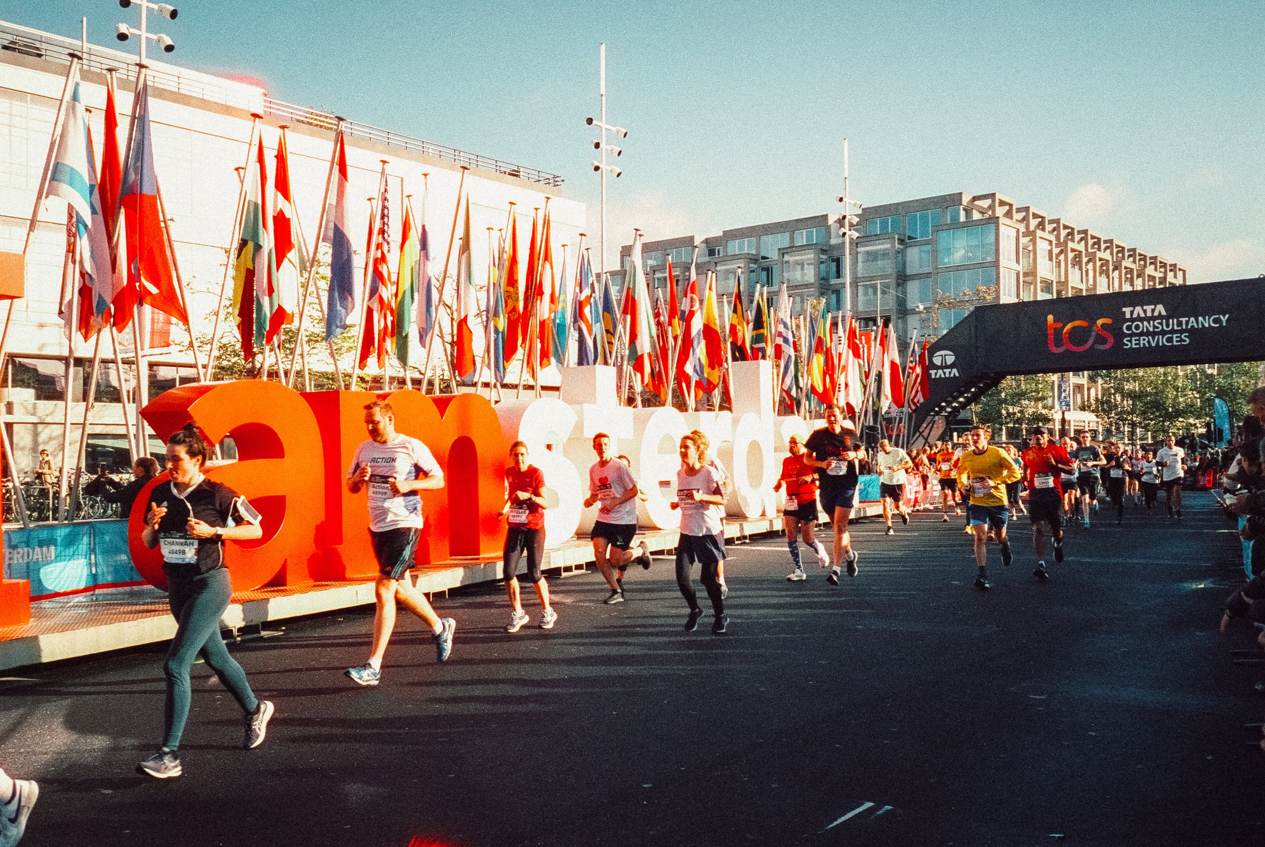 People participating in a running event, with flags and banners along the route, in front of modern buildings and a sign that reads 'Tata Consultancy Services'.