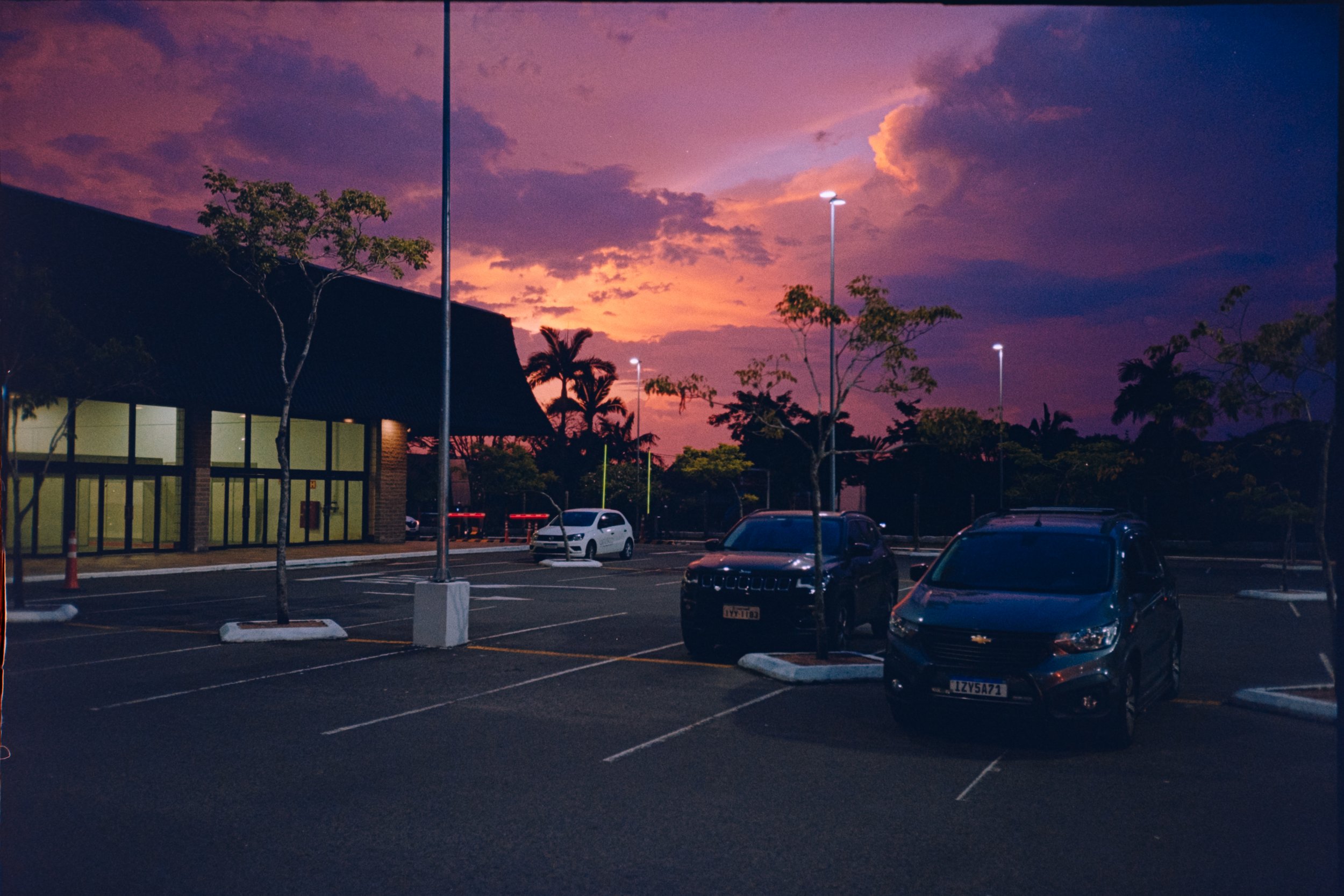 A parking lot at dusk with some cars parked, nearby trees, and a building with large windows. The sky is colorful with shades of pink, purple, and orange, and there are tall streetlights illuminating the area.
