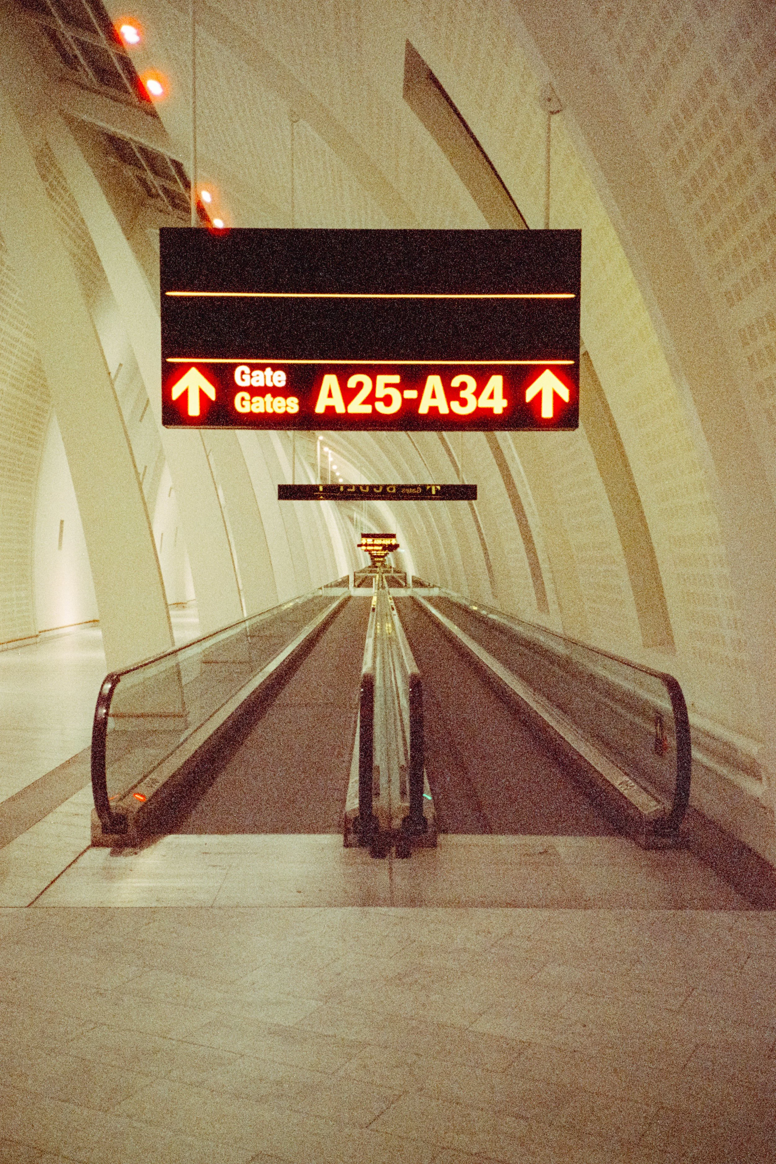An airport gate display sign showing Gates A25-A34 with upward arrows, located in an area with light-colored tile flooring and curved walls