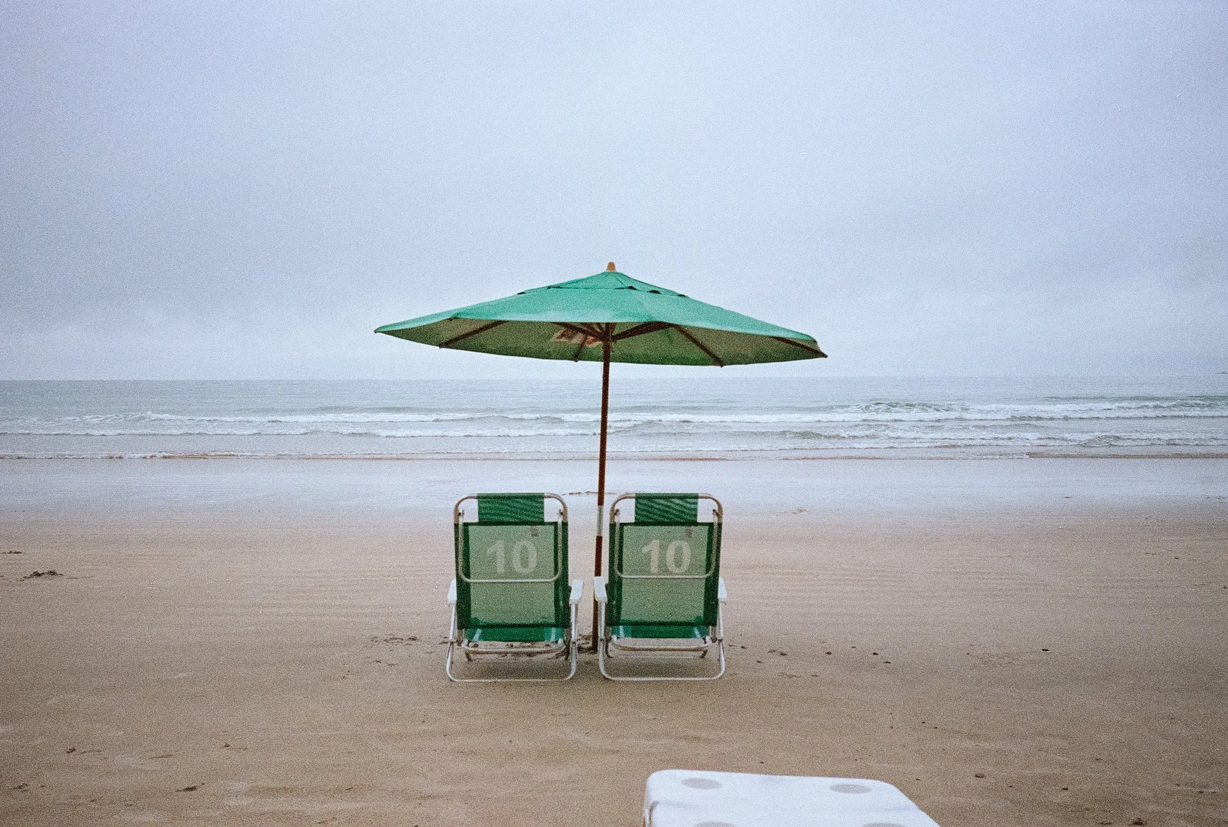 Empty beach chairs under an umbrella on the sandy beach, with the ocean and cloudy sky in the background.