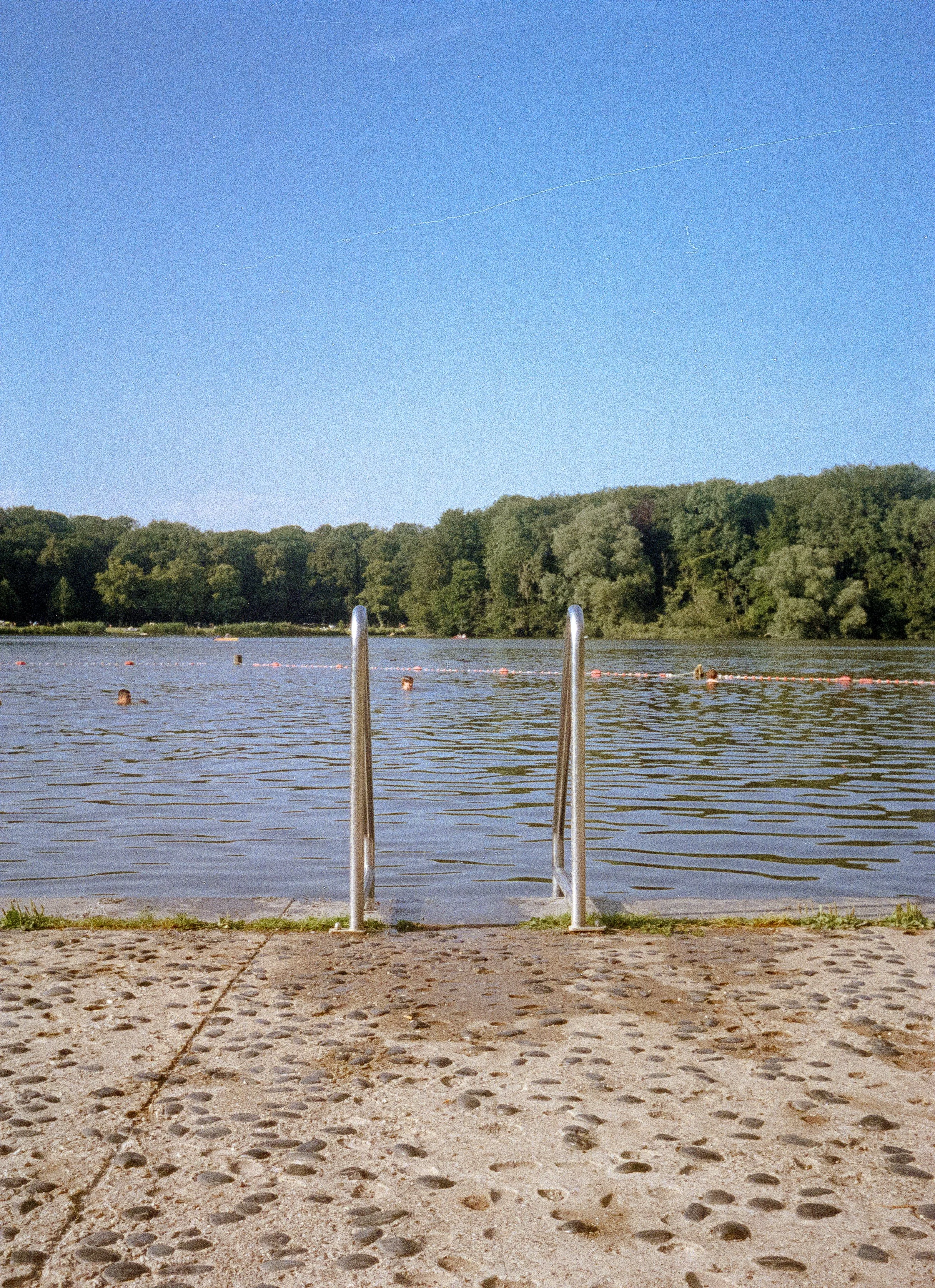 A lake with a dock and submerged ladder, swimmers in the water, green trees on the opposite shore, and a clear blue sky.