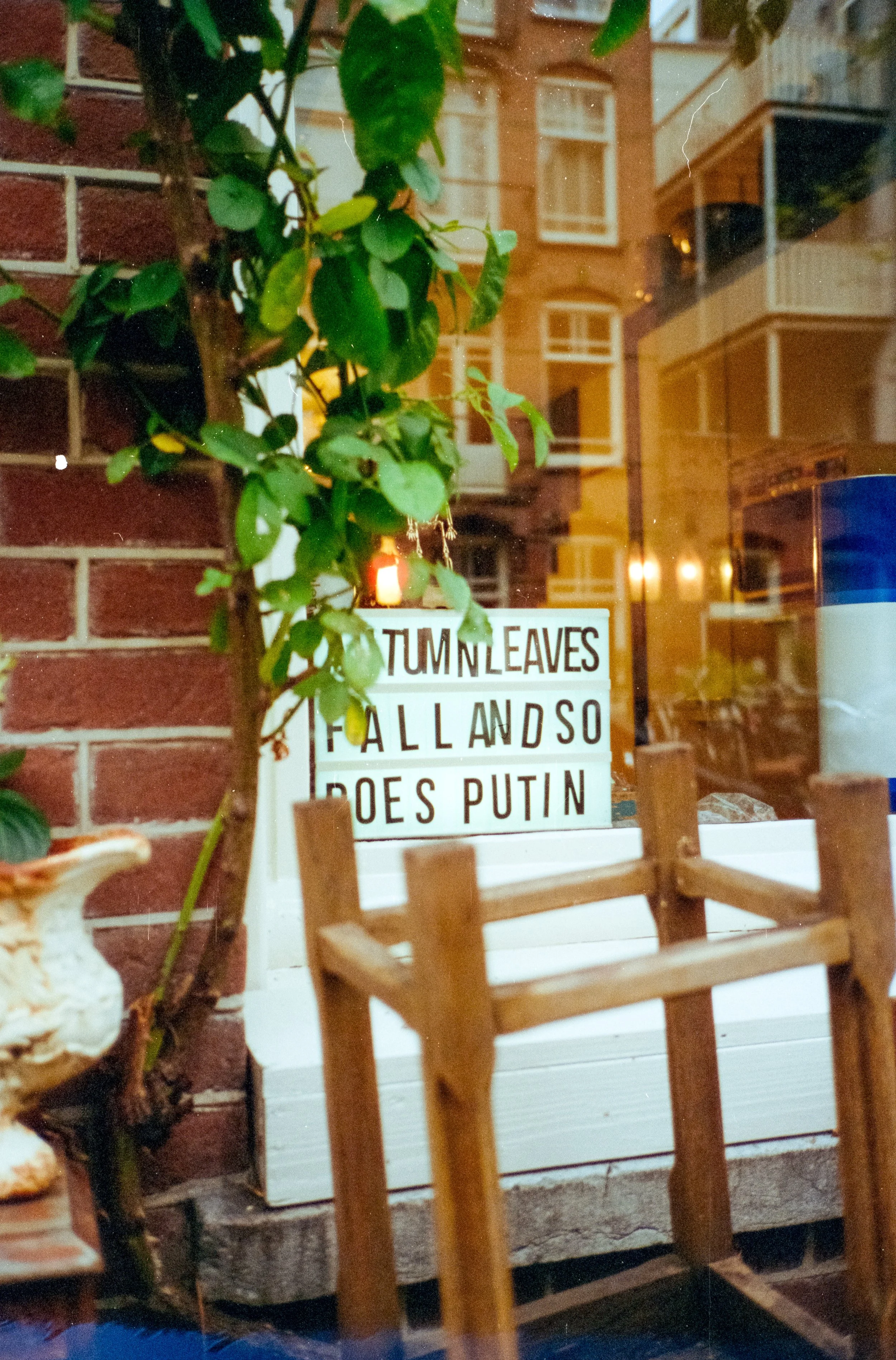 View from behind a table with a small sign that says 'Turn Leaves Fall and So Do Putin' in a restaurant or cafe. There are green leaves and a brick wall to the left, and reflections of apartment buildings can be seen in the window behind.