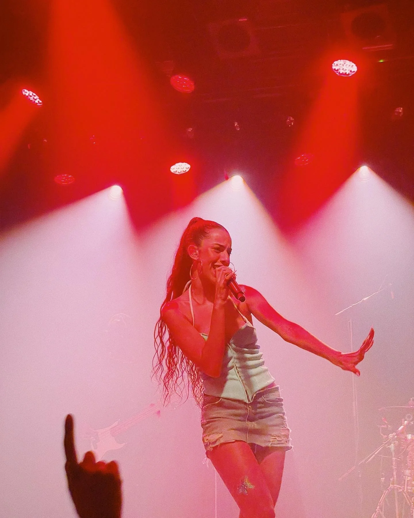 A female singer performing on stage under red lighting, with a microphone in hand and her long hair in a high ponytail.