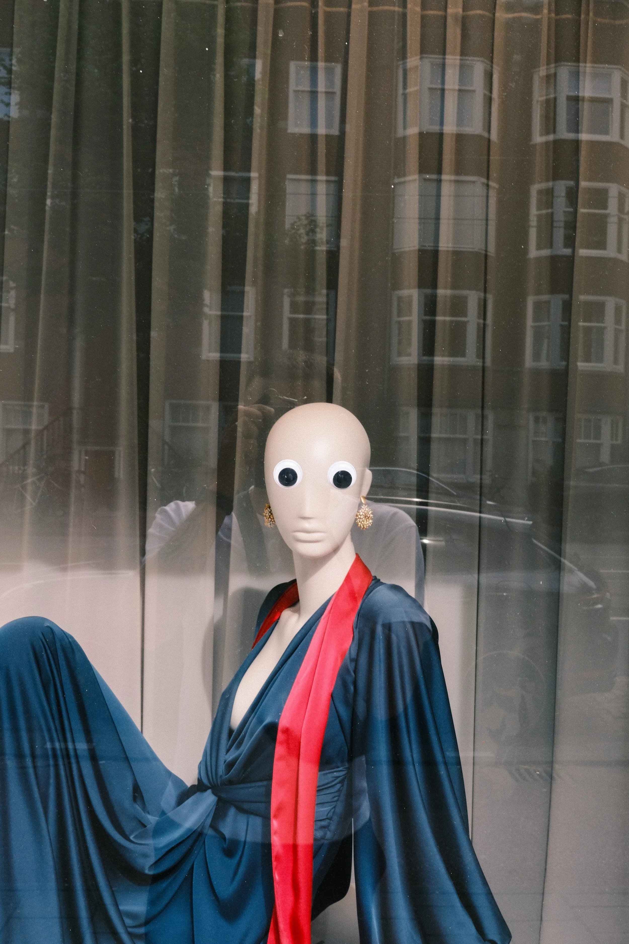 A mannequin in a store window dressed in a traditional blue kimono with a red sash and large googly eyes for a face, with a reflection of an apartment building in the background.