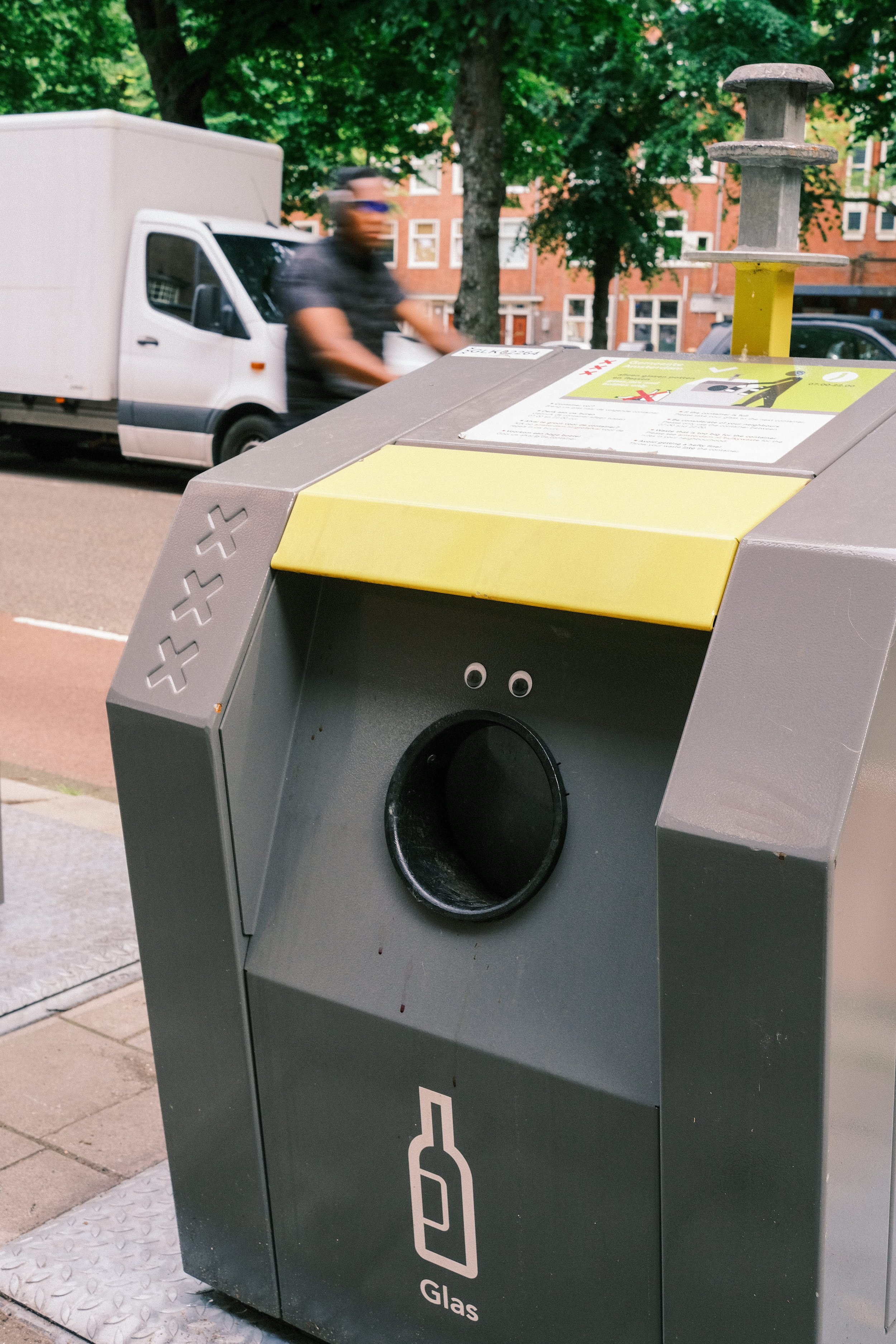 Public glass recycling bin with a white label and a modern design, situated outdoors on a sidewalk, with a blurred person and a white delivery truck in the background.
