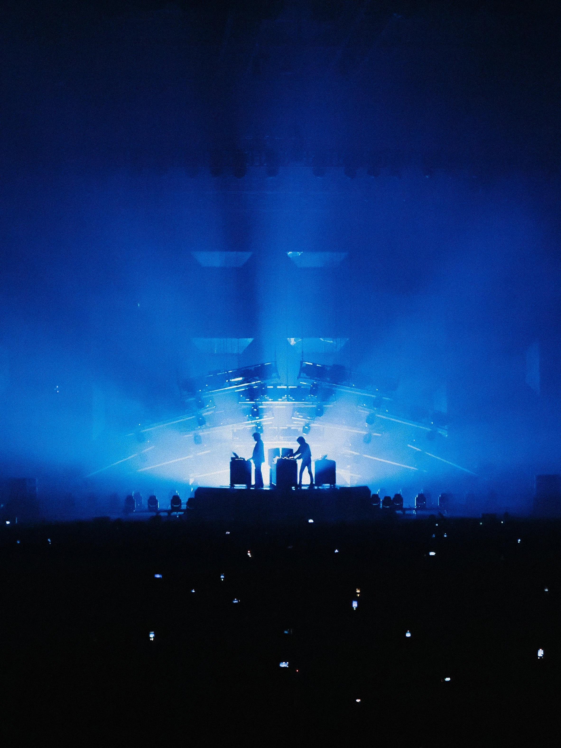 A concert stage illuminated with blue lighting, showing two silhouetted performers or DJ equipment in the foreground, with an audience and numerous small lights in the dark.