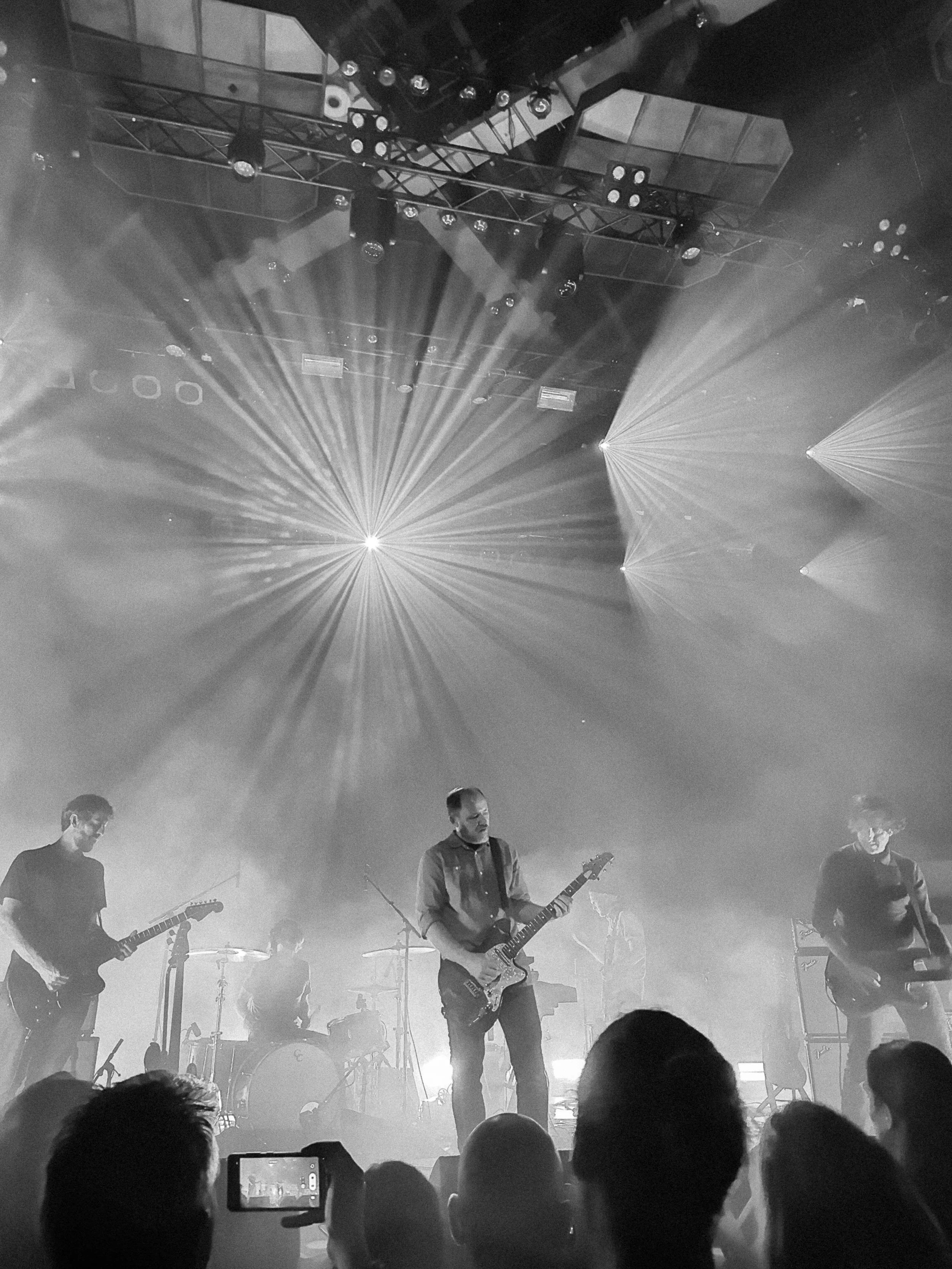 Black and white photo of a band performing on stage with four musicians playing guitars and a drummer in the background. Bright spotlights and stage lights are directed toward the performers, with the audience visible at the bottom, some recording or