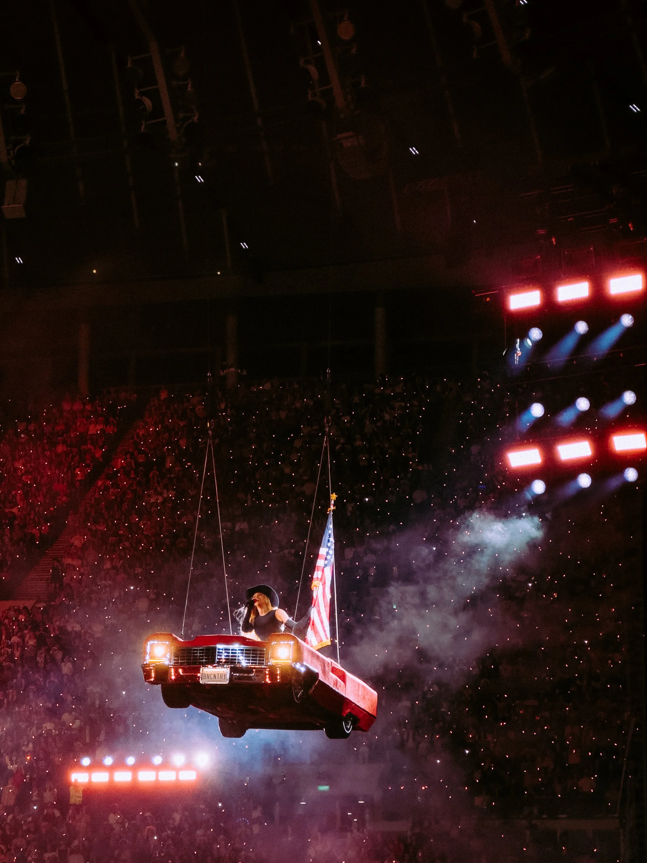 A woman is riding in a red vintage car during a stadium event, holding an American flag with lights and smoke effects around her, in front of a large crowd and a dark arena ceiling.