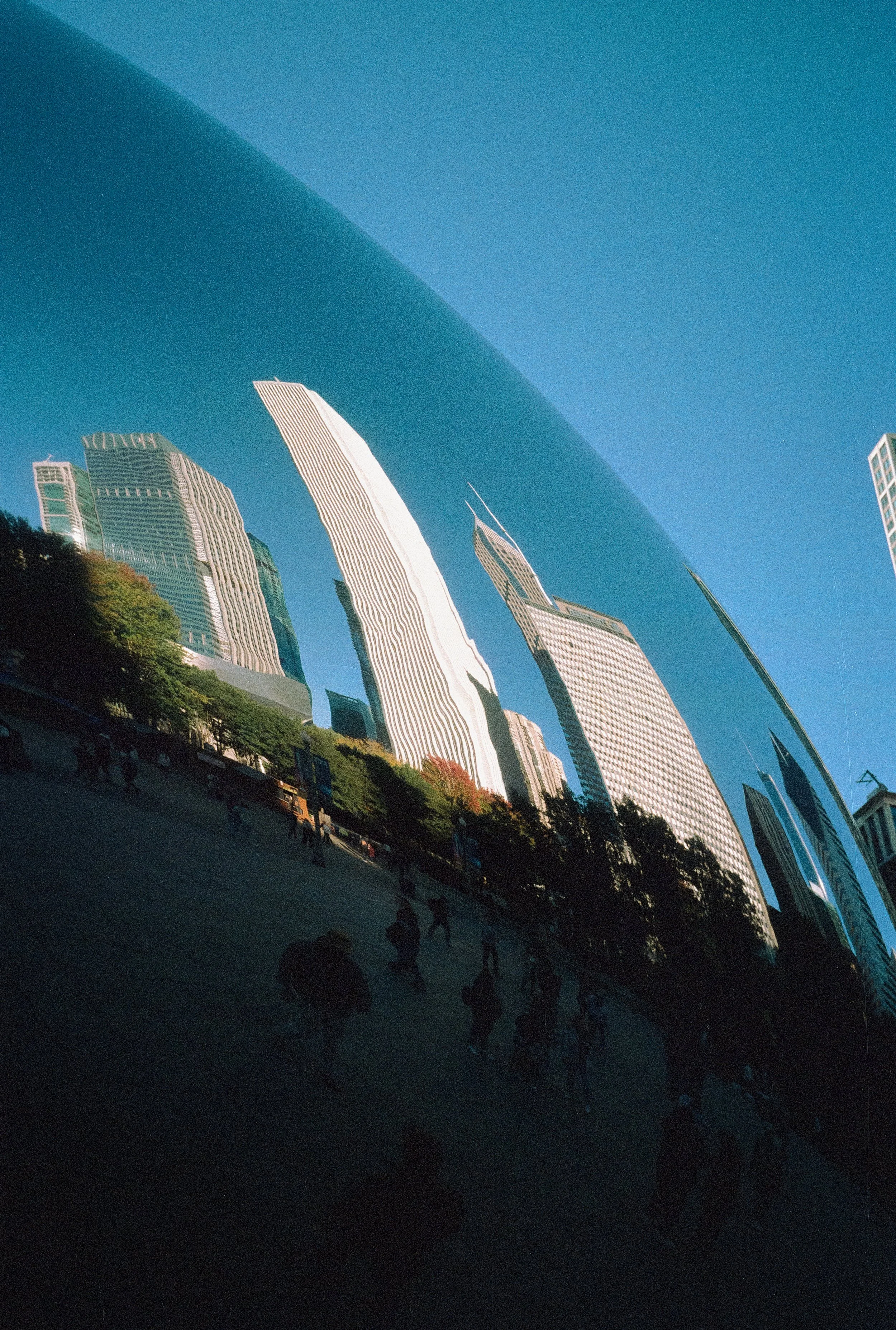 Reflected skyscrapers and blue sky with few clouds, seen in the shiny surface of a large building, with a small silhouette of people at the bottom.