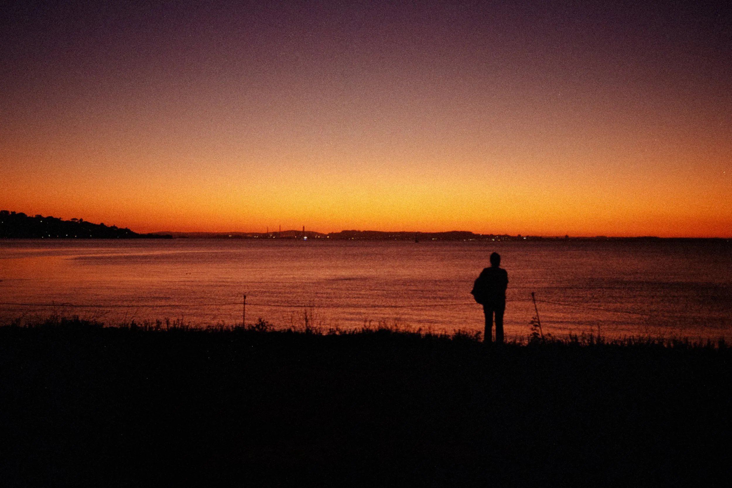 A person standing by the water at sunset or sunrise, with a colorful sky and distant bridge in the background.