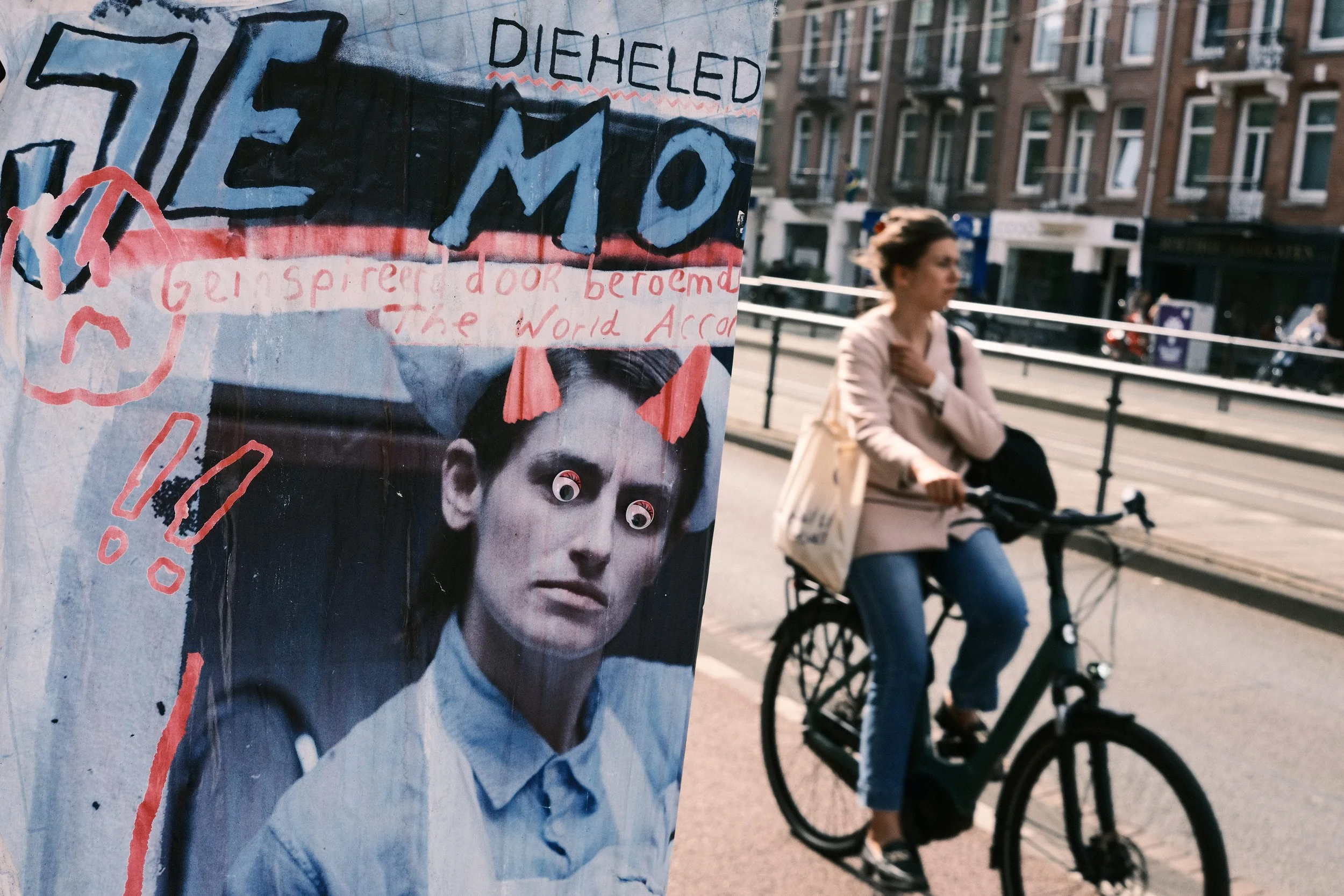 A protest poster with a surreal image of a woman with googly eyes, and handwritten text including the words 'DIEHELD' and 'GEINSPIREER door beroemd' in an urban setting with a woman on a bicycle in the background.