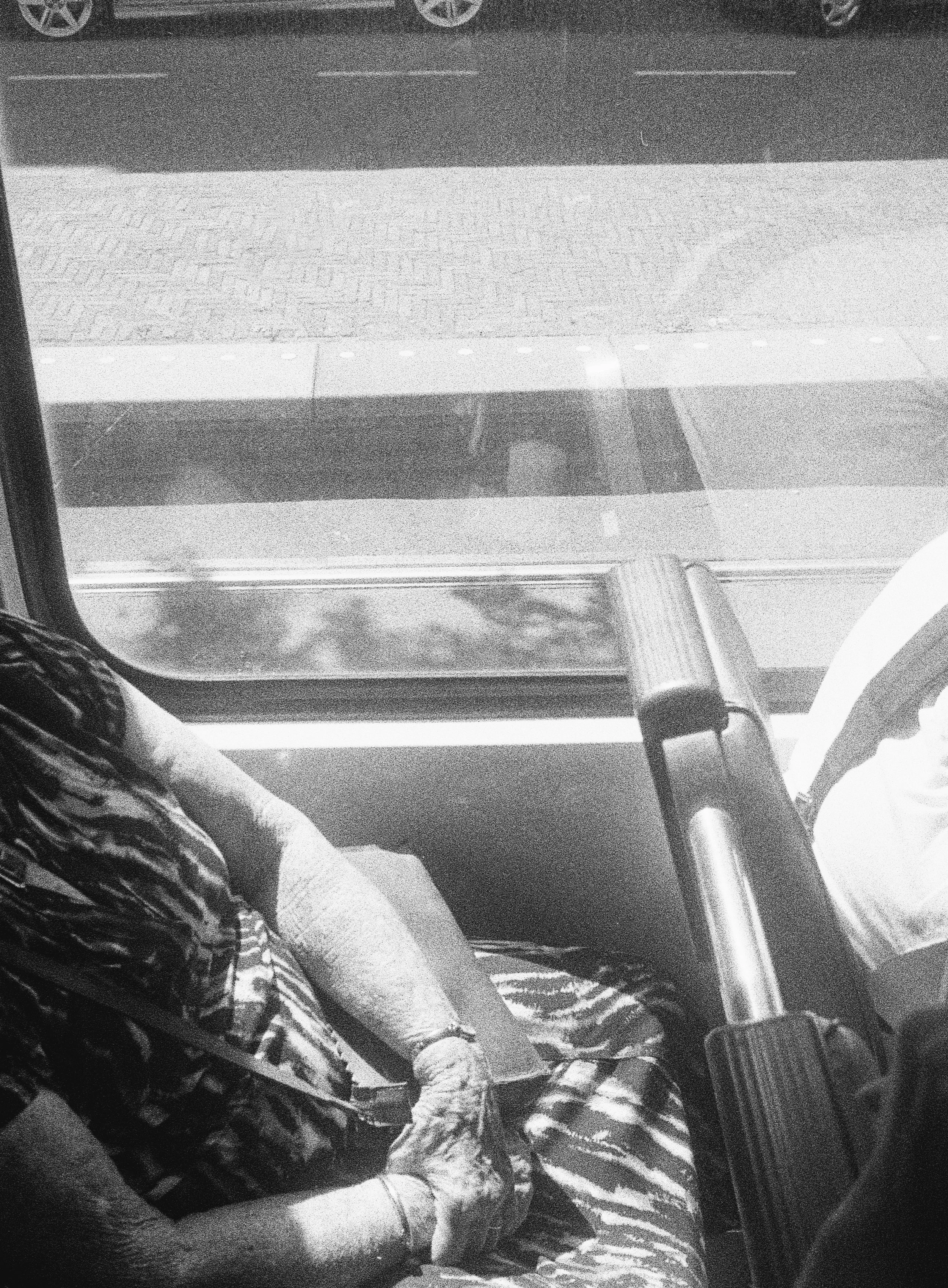 A person wearing striped pants and a patterned top, sitting on a seat with their left arm resting on their lap, behind a glass window, with a parking lot visible in the background.