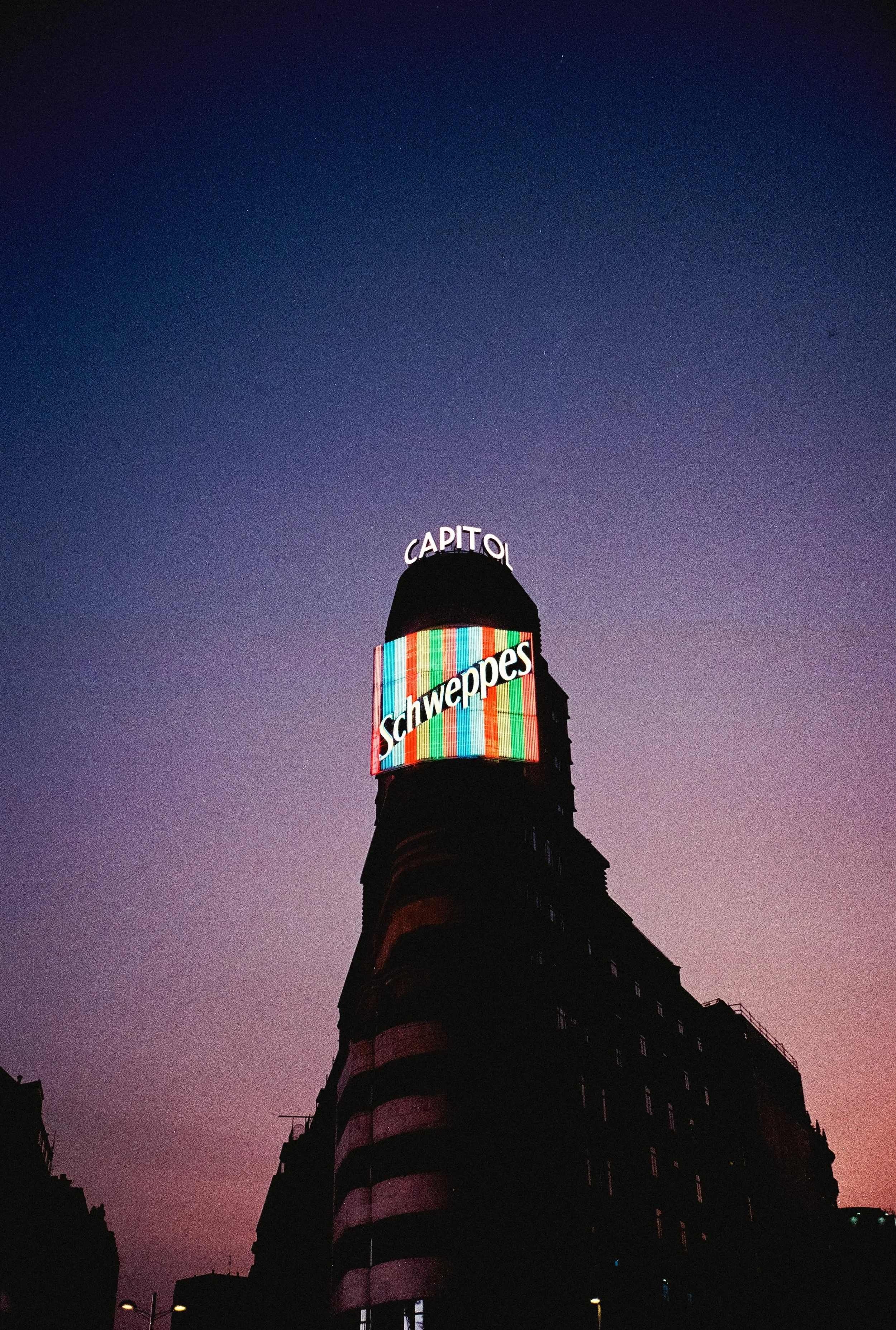 A tall building in the city at dusk with a colorful electronic billboard and a lit-up sign reading 'CAPITOL' near the top.