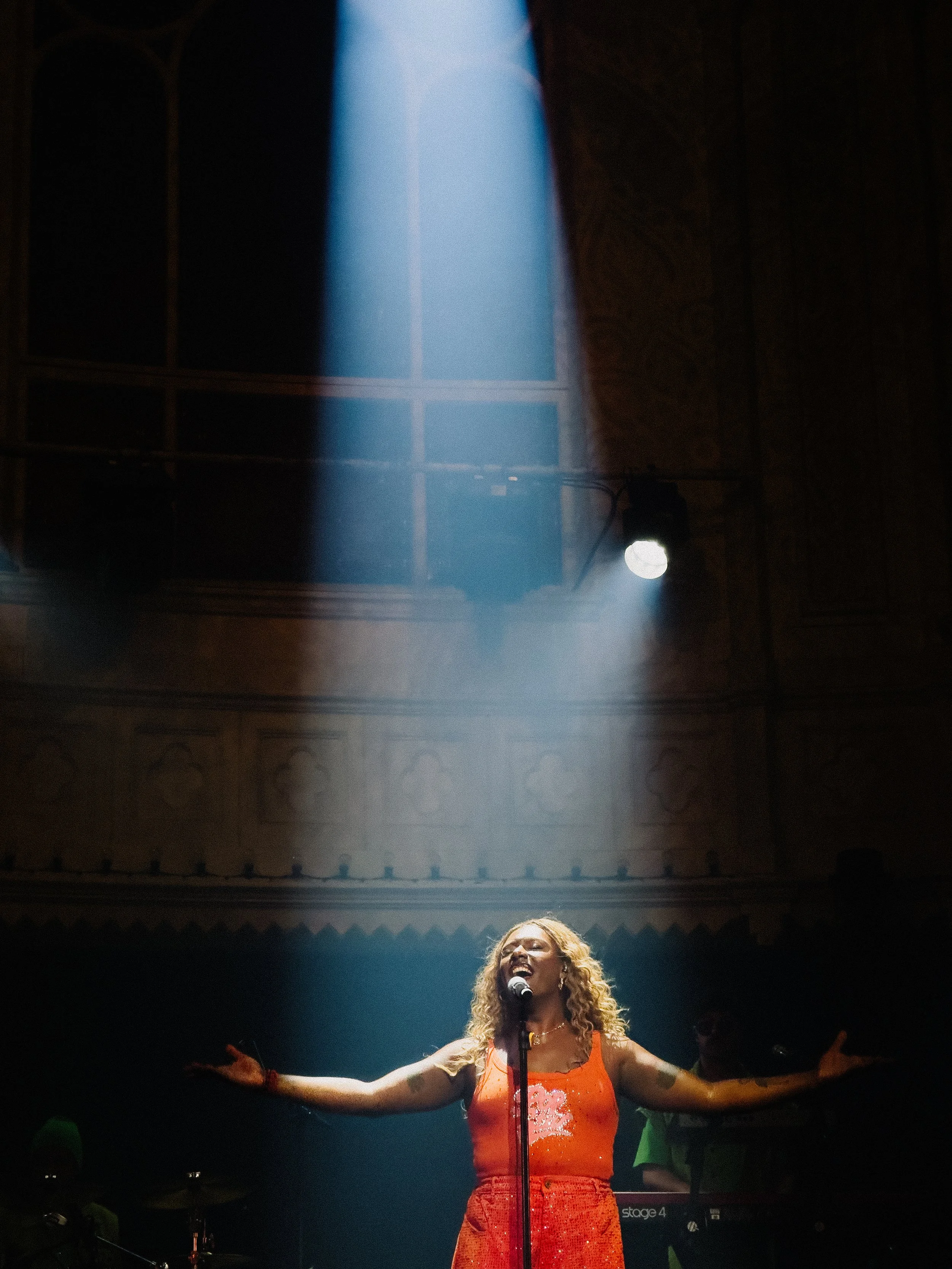 A woman with curly hair singing passionately on stage, wearing a red, sparkly dress with outstretched arms, illuminated by stage lights in a dark venue.