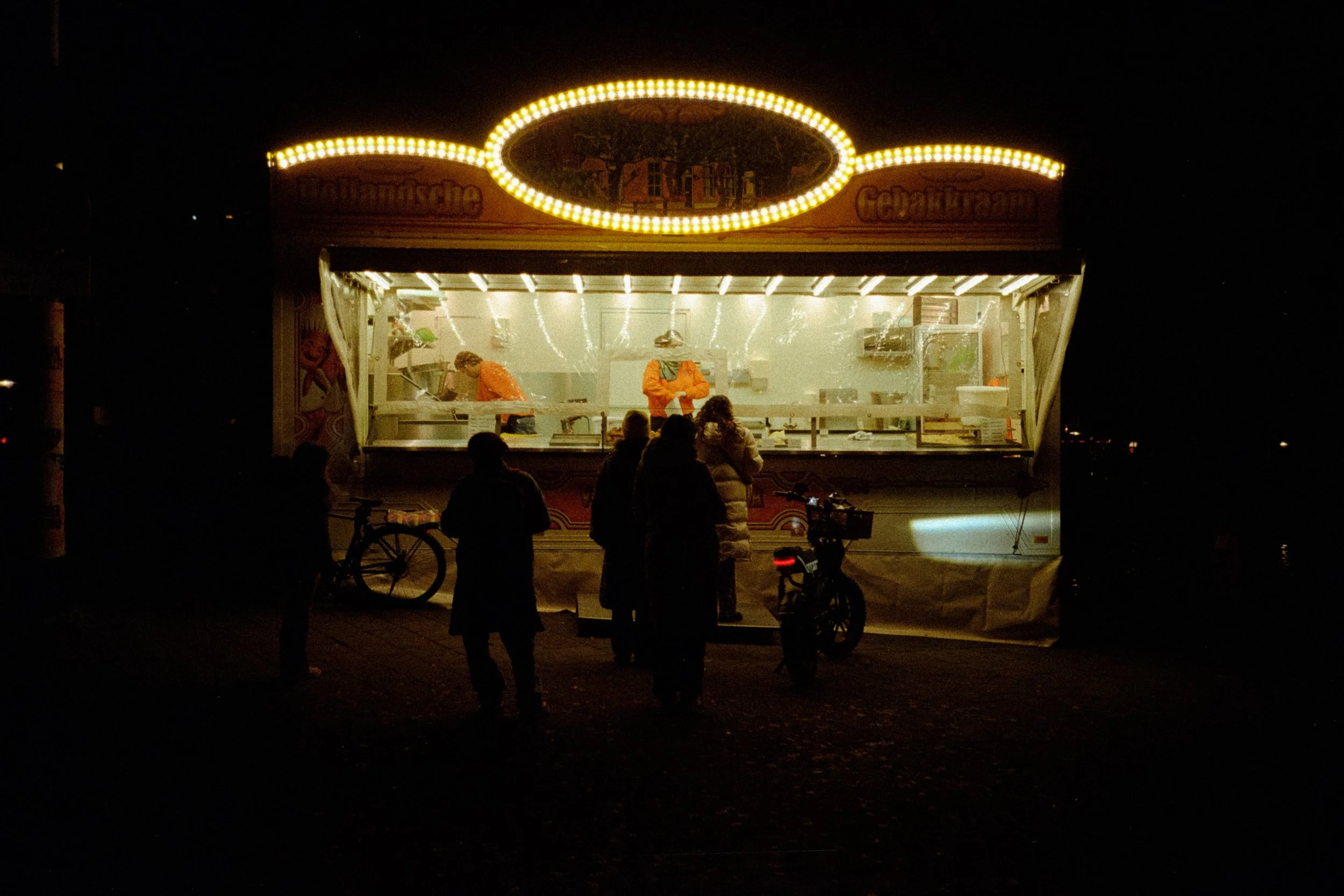 A brightly lit food stand at night with people gathered in front, some on bicycles, with a dark background.