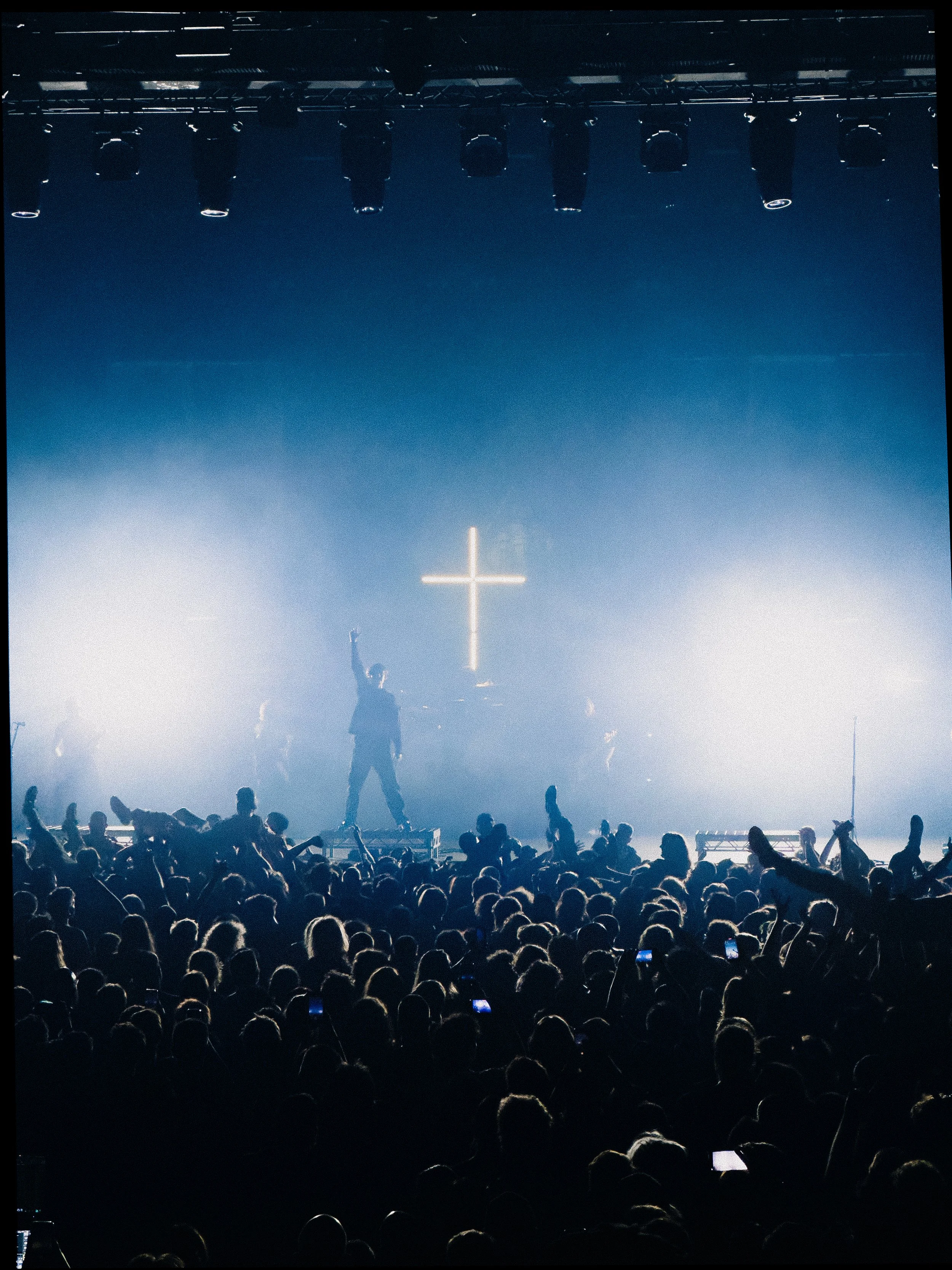 Concert crowd facing stage with a singer and band, with a large illuminated cross in the background and multiple stage lights overhead.