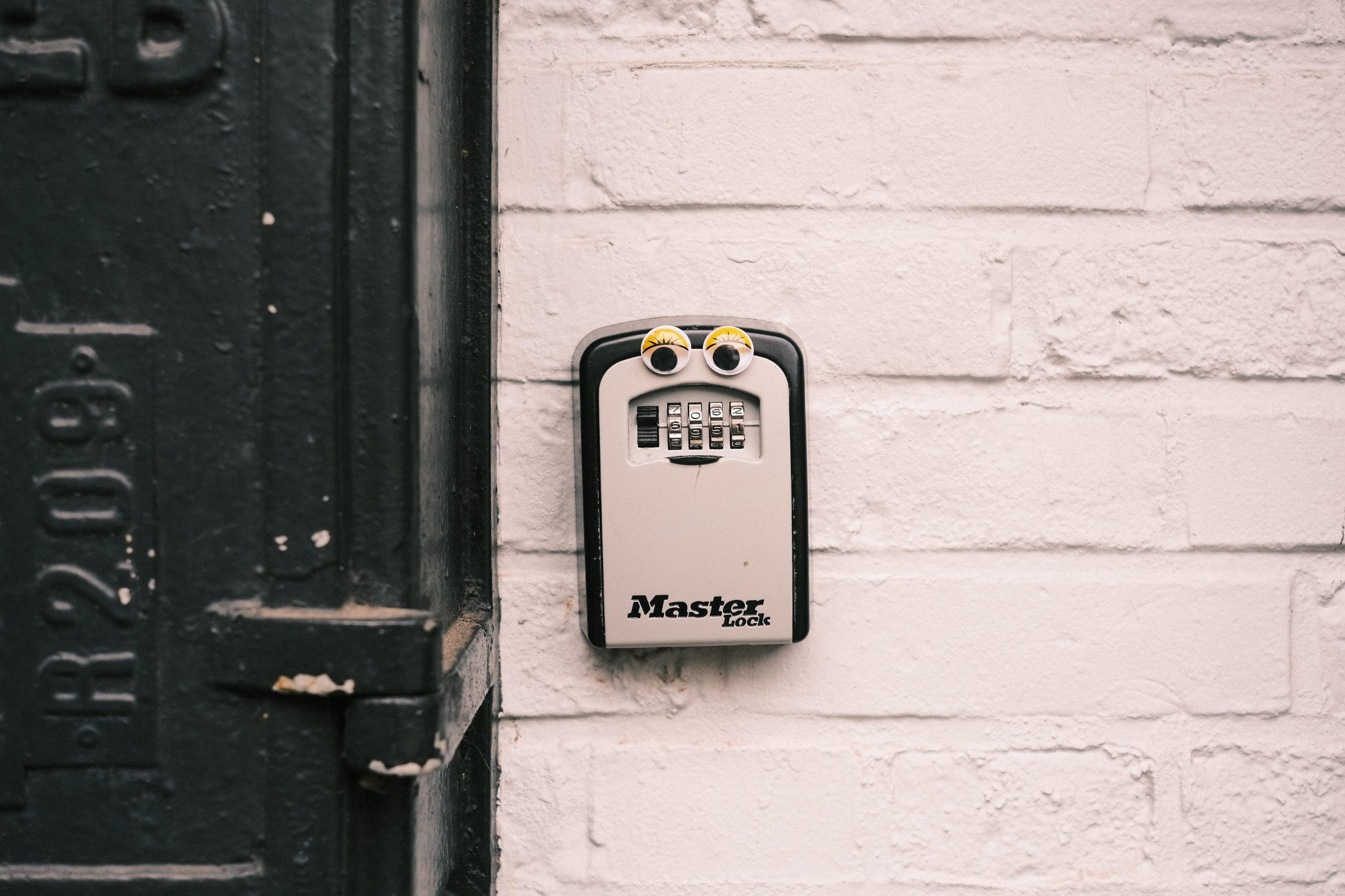 A doorbell with two googly eyes attached to it, mounted next to a black metal door frame and a white brick wall.