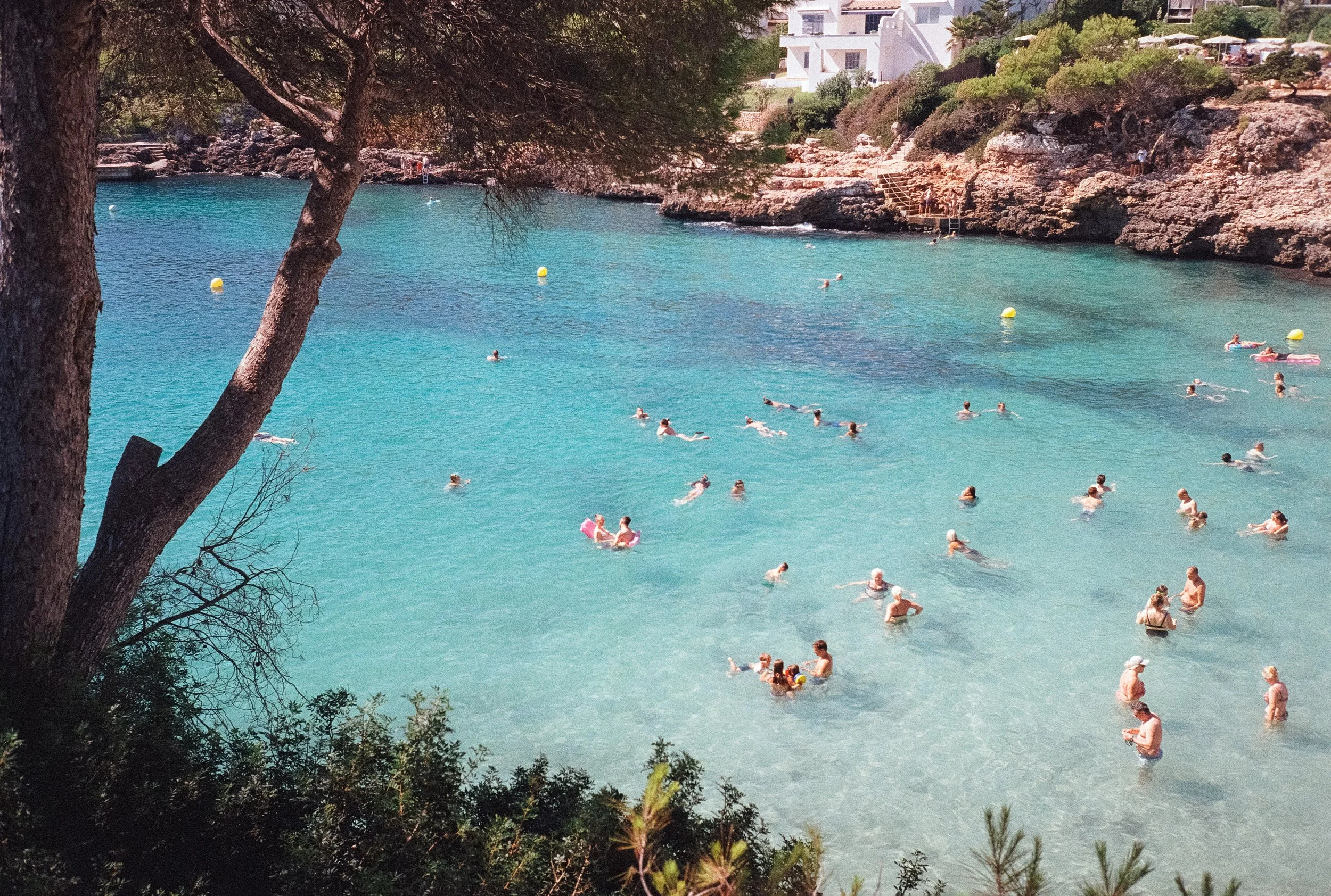 People swimming and relaxing in a clear, turquoise body of water near rocky shoreline, framed by trees and houses in the background during daytime.