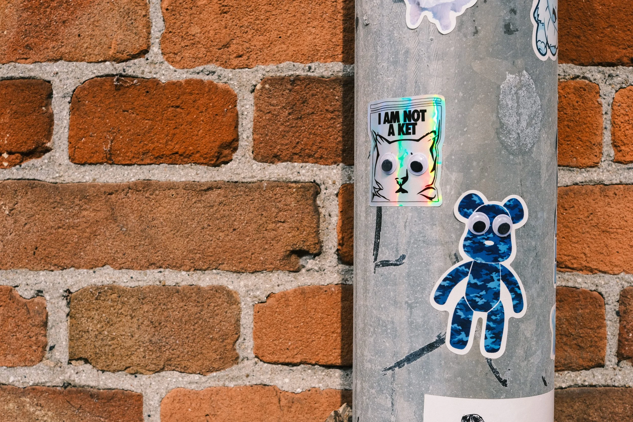 Street art stickers on a metal pole next to a red brick wall. One sticker says "I am not a ket" with a drawing of a cat; another sticker appears to be a cartoonish animal with googly eyes.