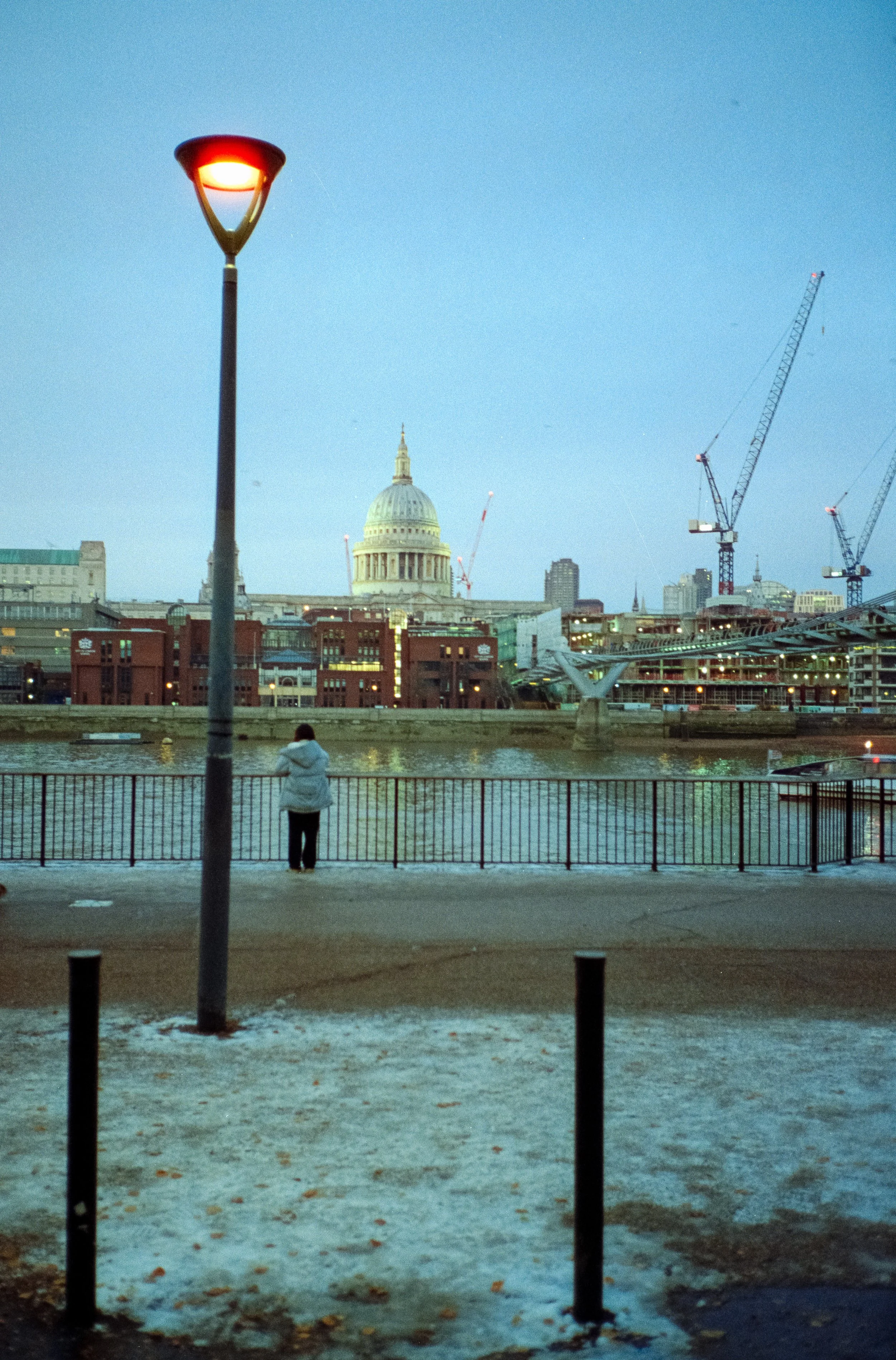 A woman in a white jacket standing near a fence along the water, with city buildings, cranes, and the U.S. Capitol in the background, during dusk or early evening.