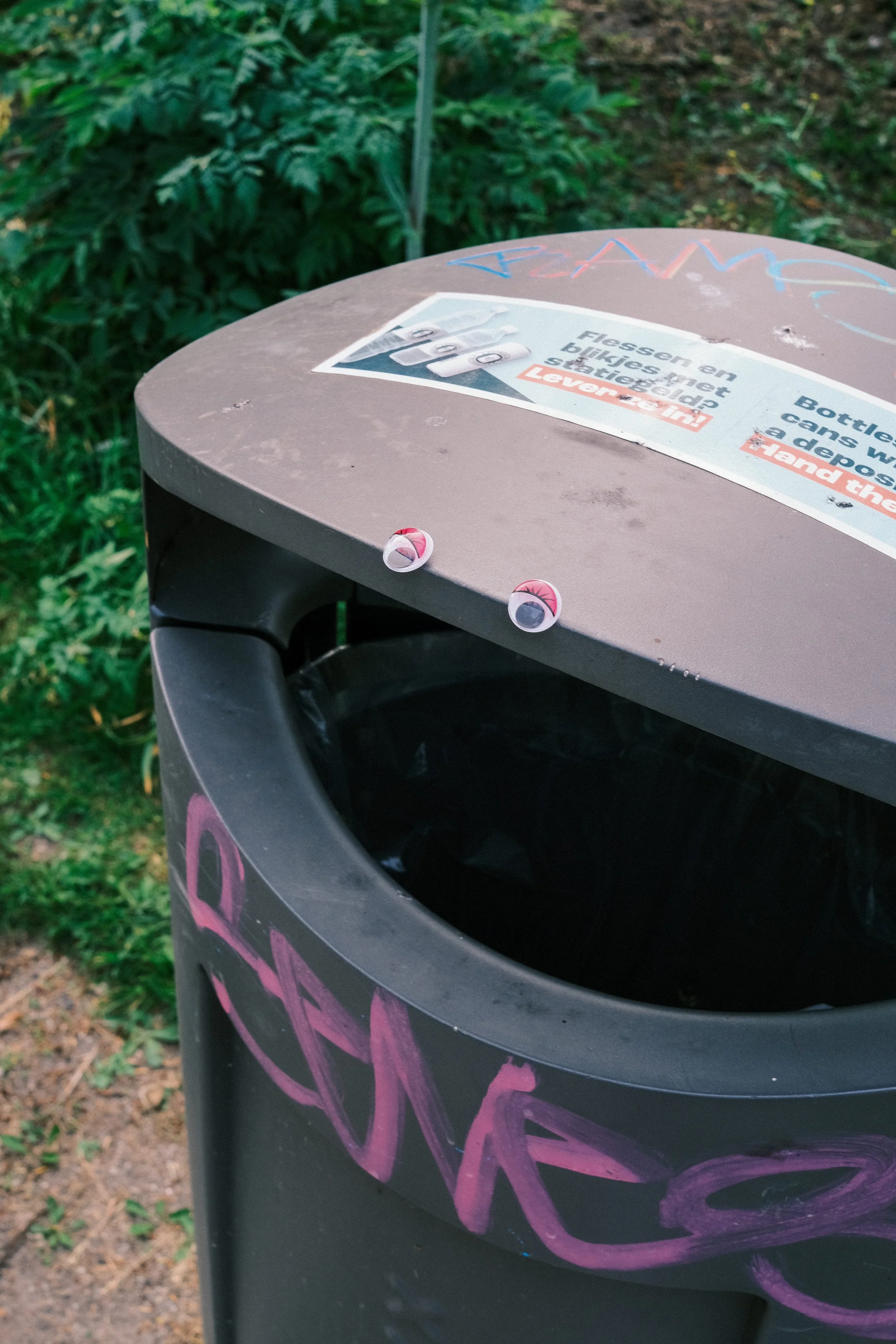 A black trash bin decorated with pink graffiti and stickers, positioned outdoors with green vegetation in the background.