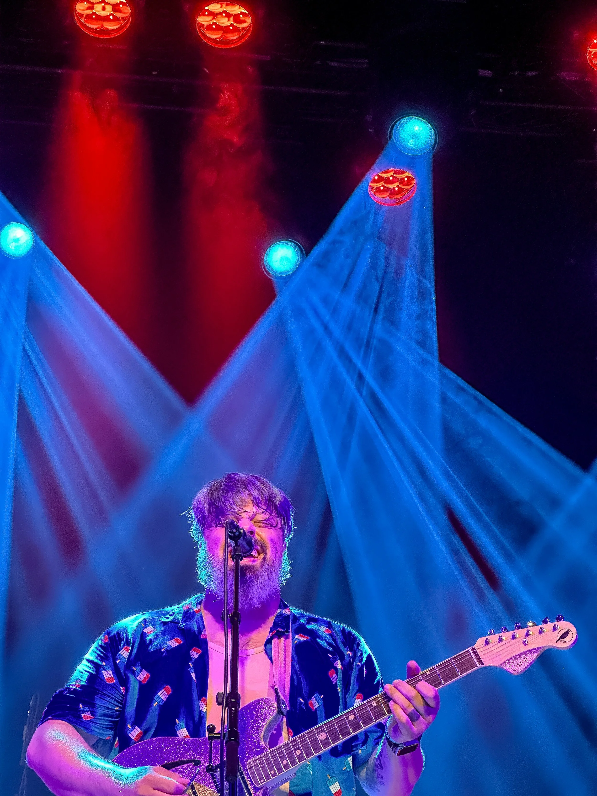 Man with glasses and beard playing guitar and singing into a microphone on stage under blue and red lights.