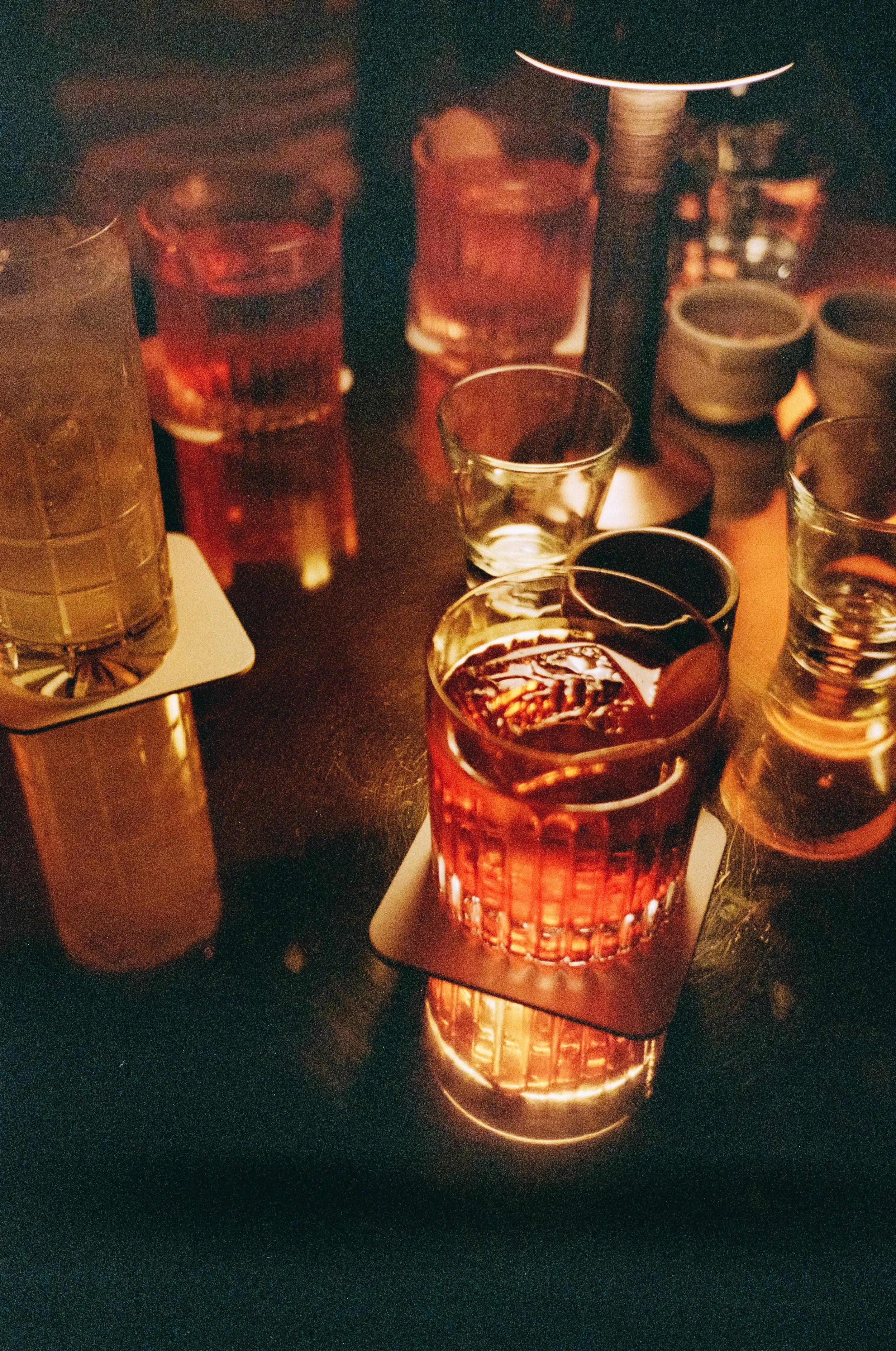 A collection of glasses filled with dark beverages, illuminated with warm lighting, on a dark table at a bar or restaurant.