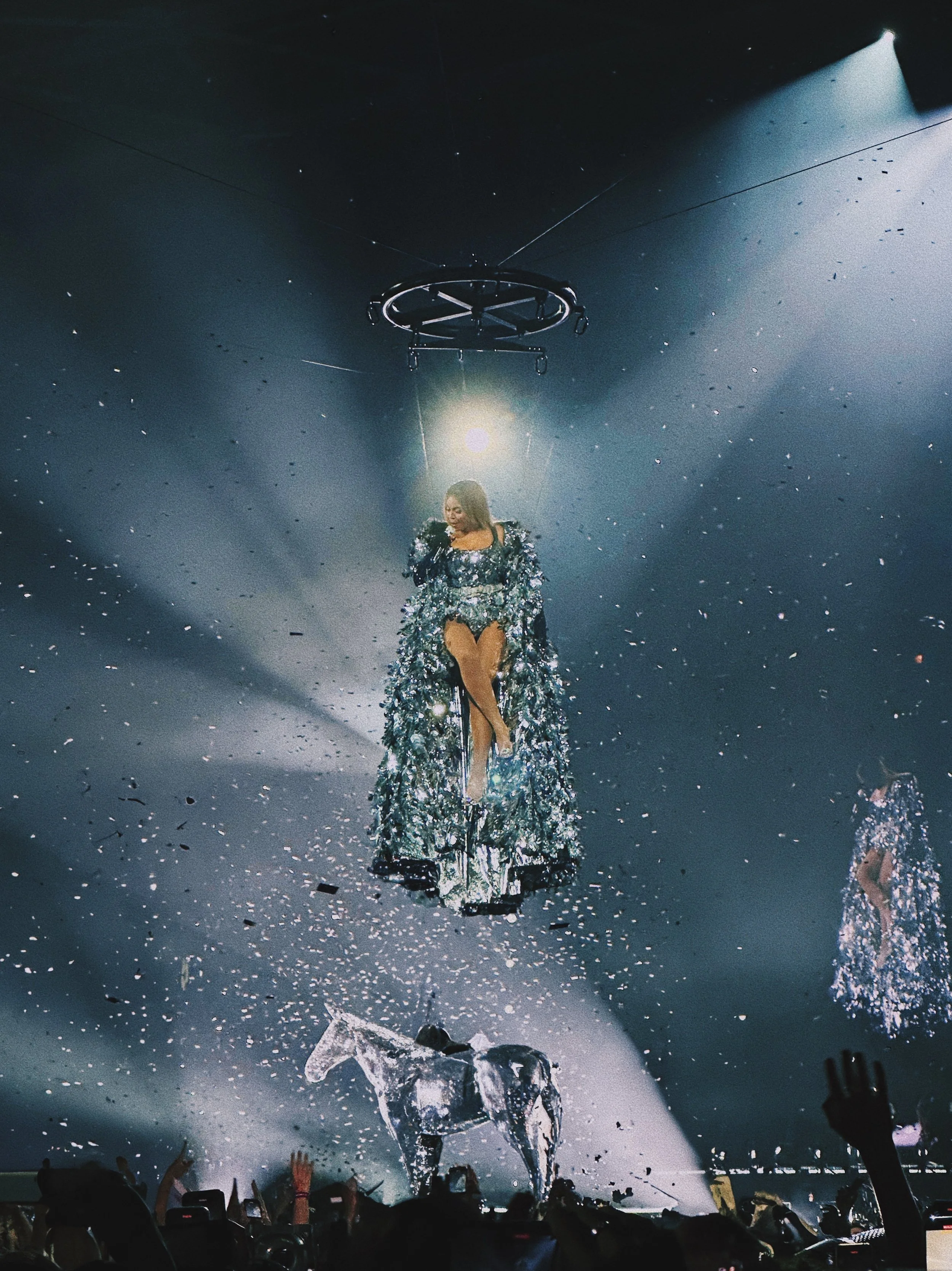 A performer on stage during a concert, with bright spotlights, confetti in the air, and audience members raising their hands.