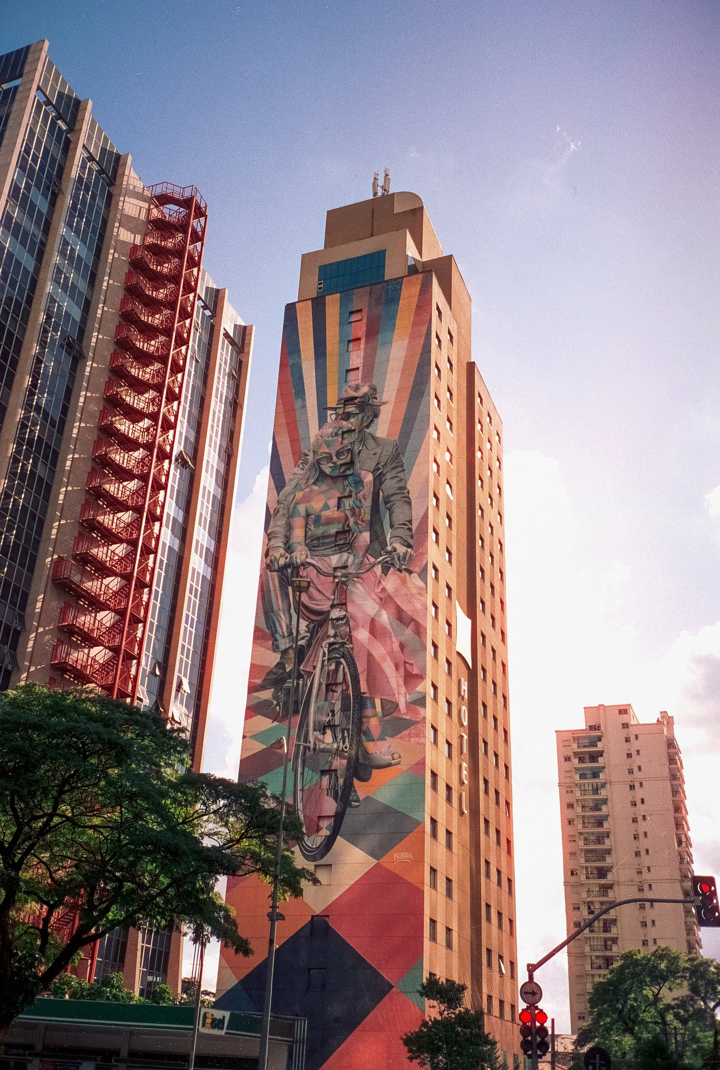 Tall building with colorful geometric mural featuring a man and woman riding a bicycle, surrounded by other tall buildings and green trees, with a blue sky overhead.