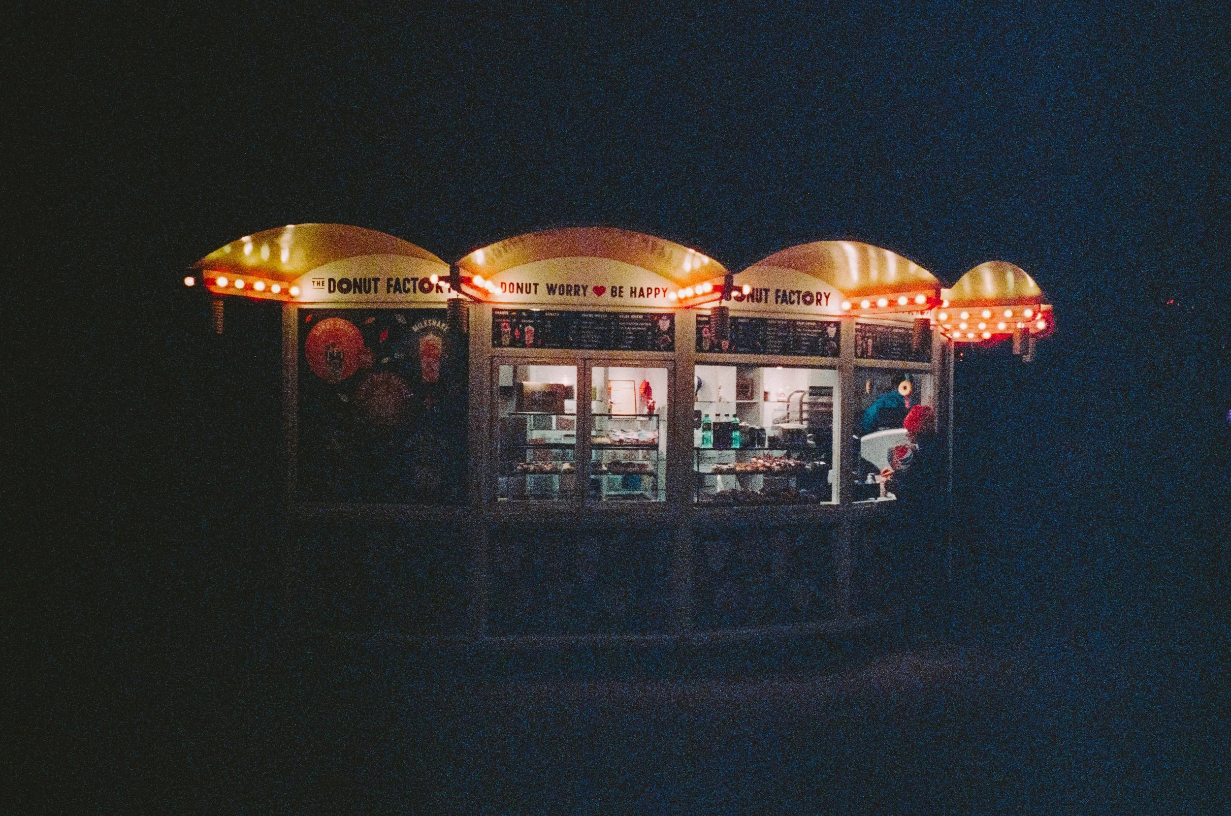 Nighttime photo of a small illuminated bakery stall called The Donut Factory, with red and white lights outlining the display area.