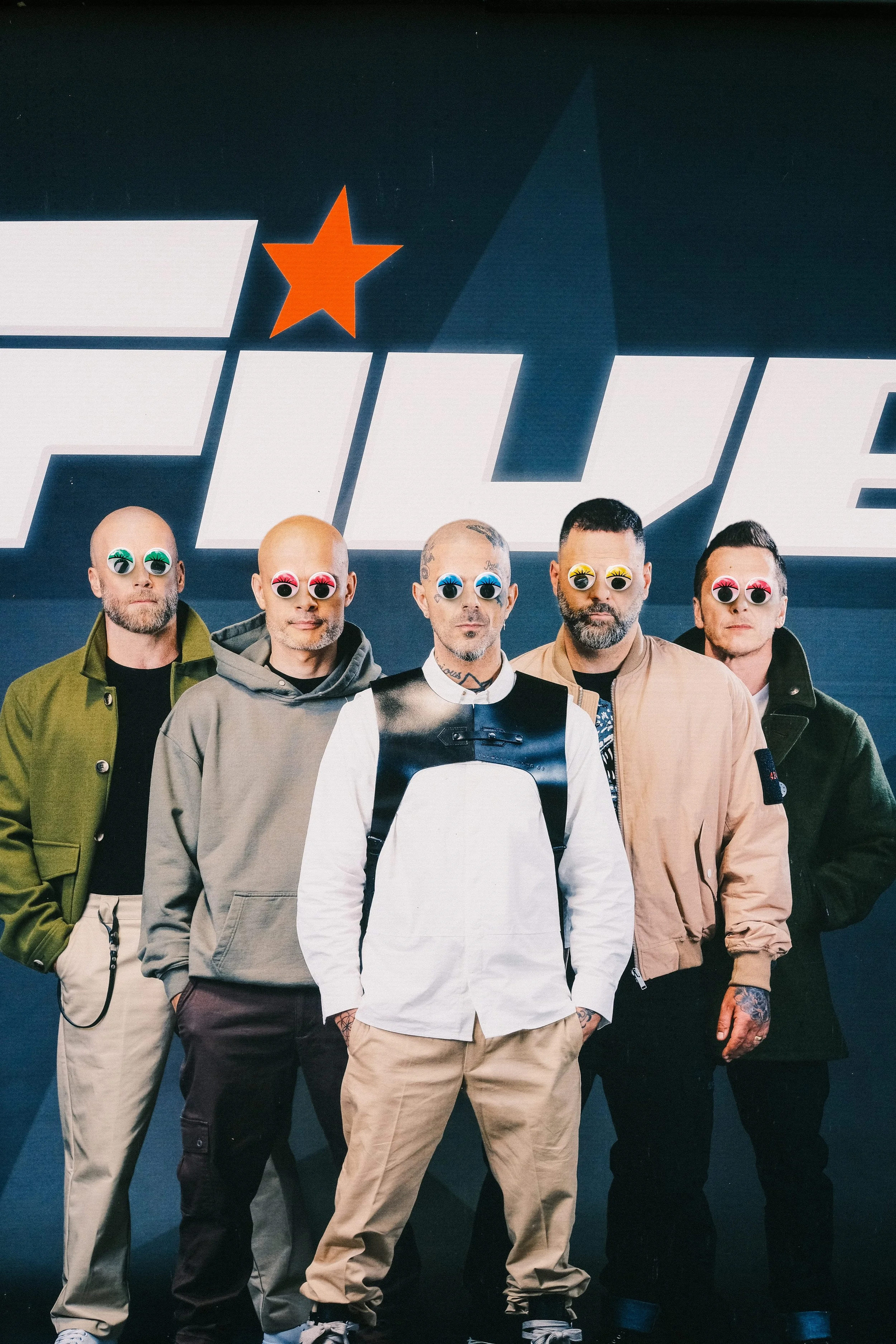 Five men standing in front of a dark background with a logo, all wearing colorful googly eye glasses. They are dressed in casual streetwear, with some tattoos visible.