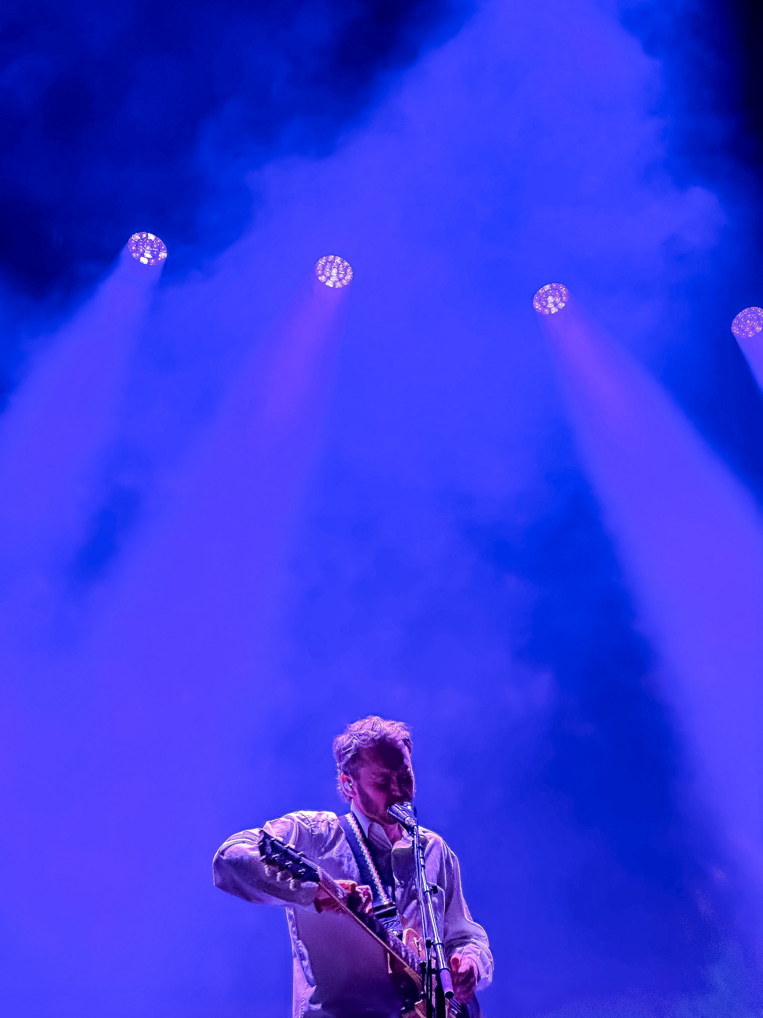 A musician with light hair playing a guitar and singing into a microphone on a stage with blue lighting.
