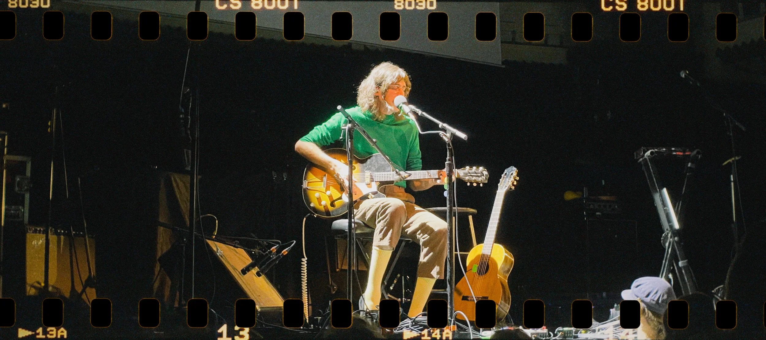 A musician with long gray hair and glasses, wearing a green shirt, is sitting on a stool on stage, playing an acoustic guitar, and singing into a microphone. There are other guitars and music equipment around him, with a dark background.