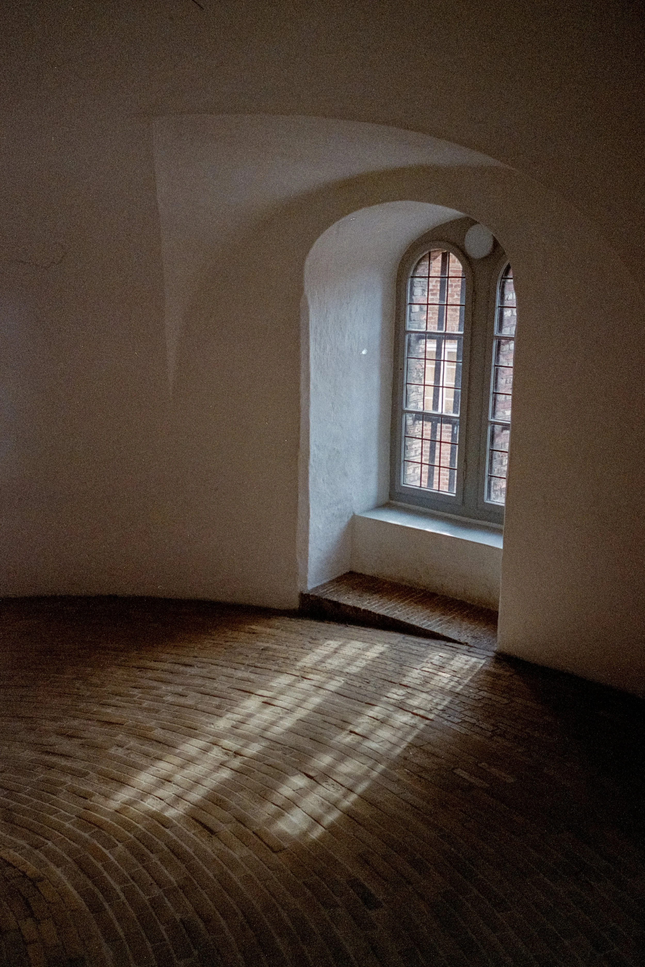A curved wooden floor next to a textured white wall and a window with brick exterior visible outside.