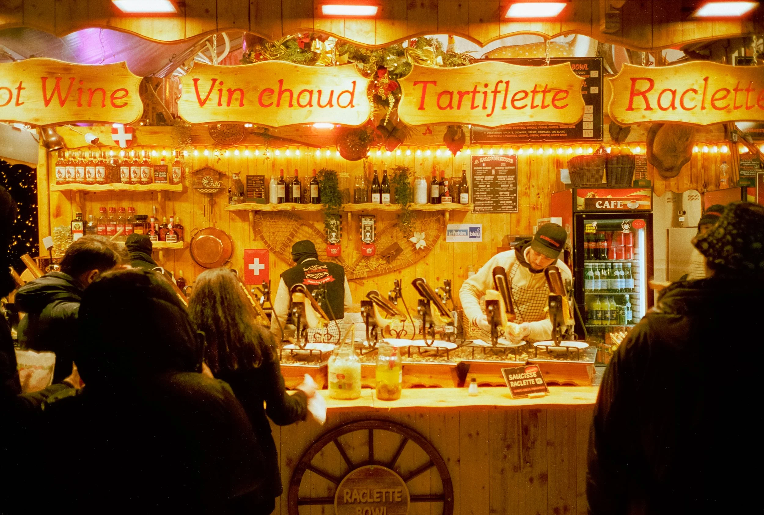 A festive food stand decorated with Christmas garlands, selling wines, tartiflette, raclette, and hot wine. Several people are gathered around, and the stand has illuminated signs with French words for various dishes and drinks.