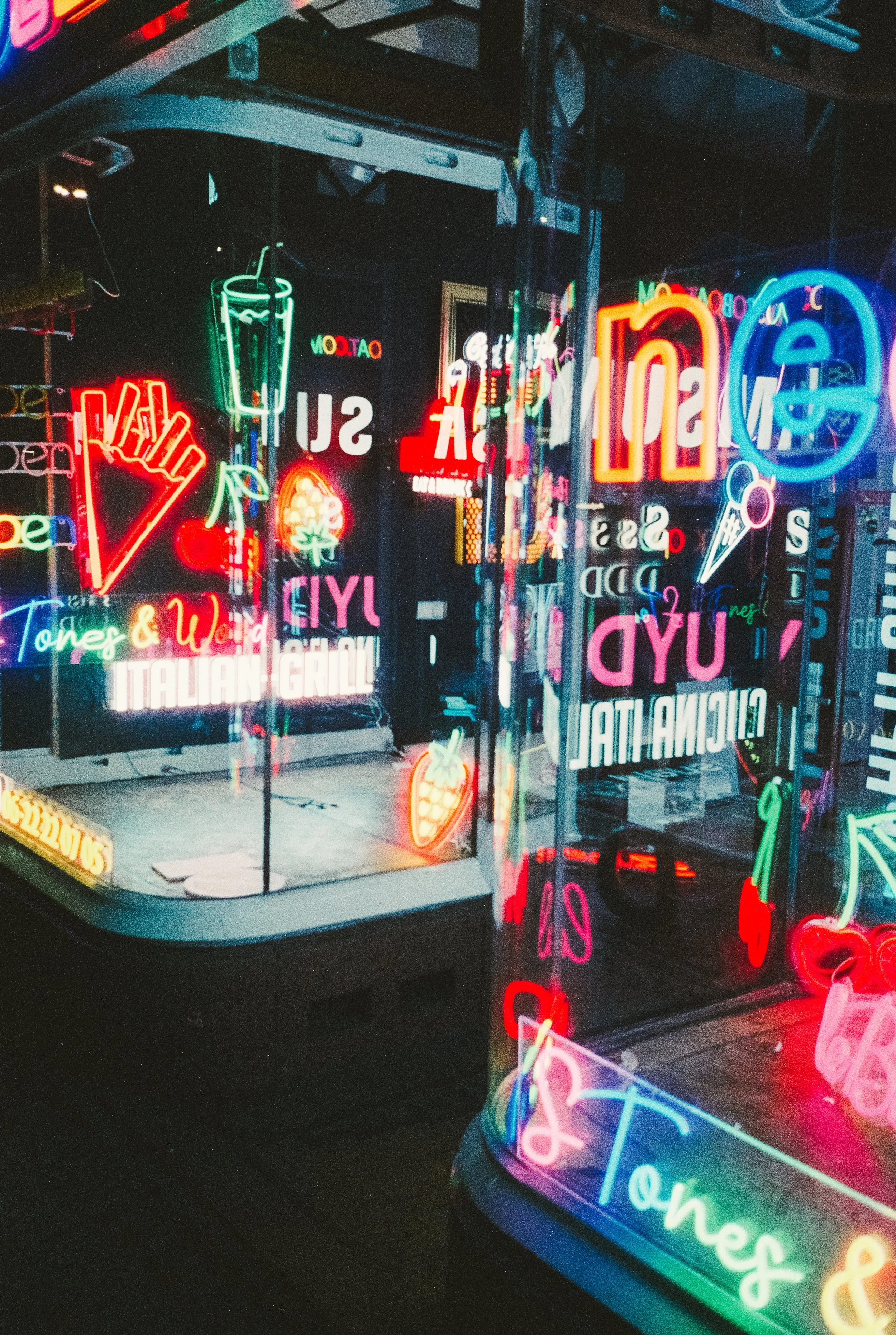 Colorful neon signs for a bar or restaurant, including a martini glass and fruit, with reflections on glass surfaces.