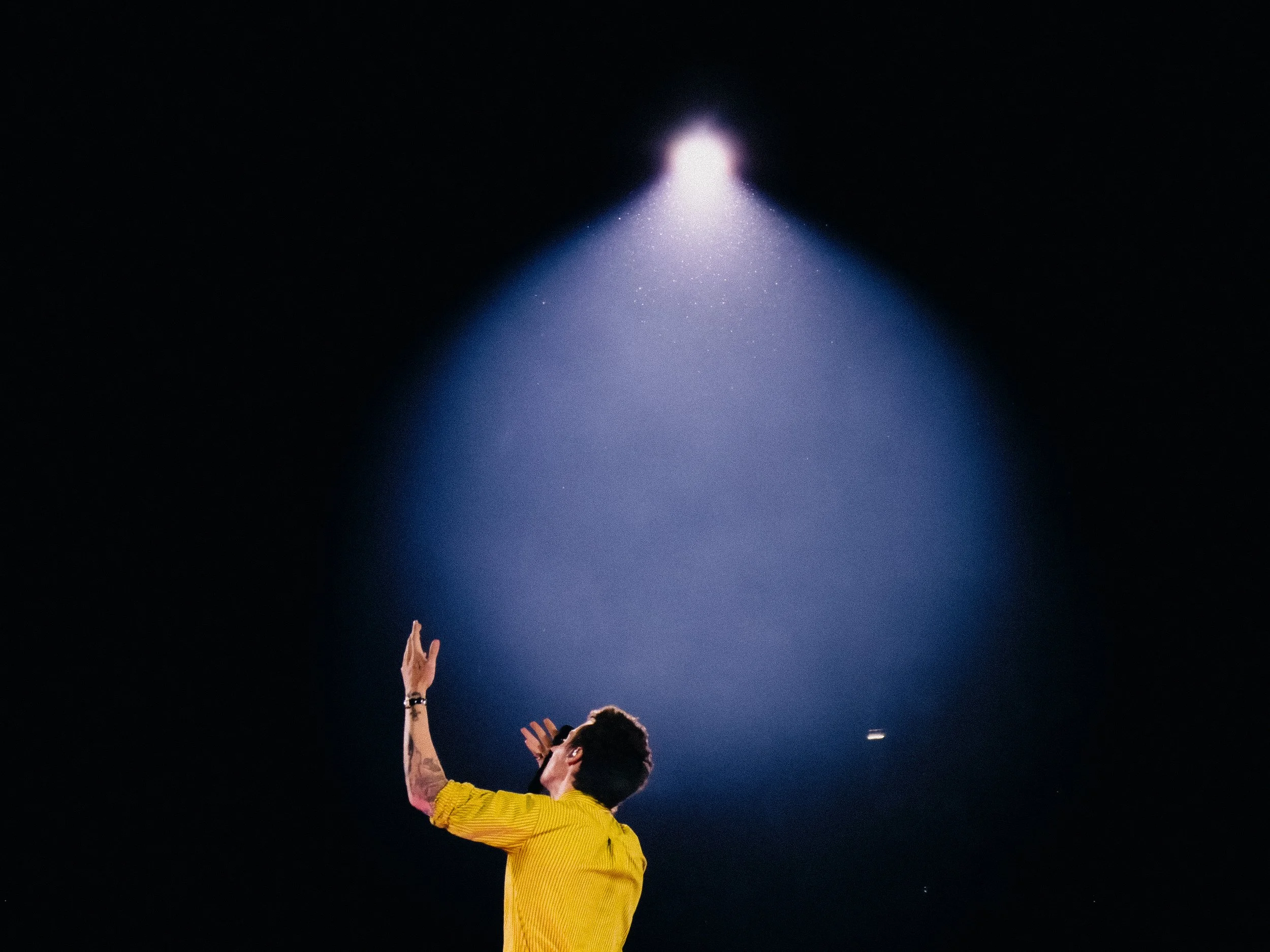 A person in a yellow jacket reaching up towards the sky at night, with their hand open and fingers spread, beneath a spotlight or bright light source.