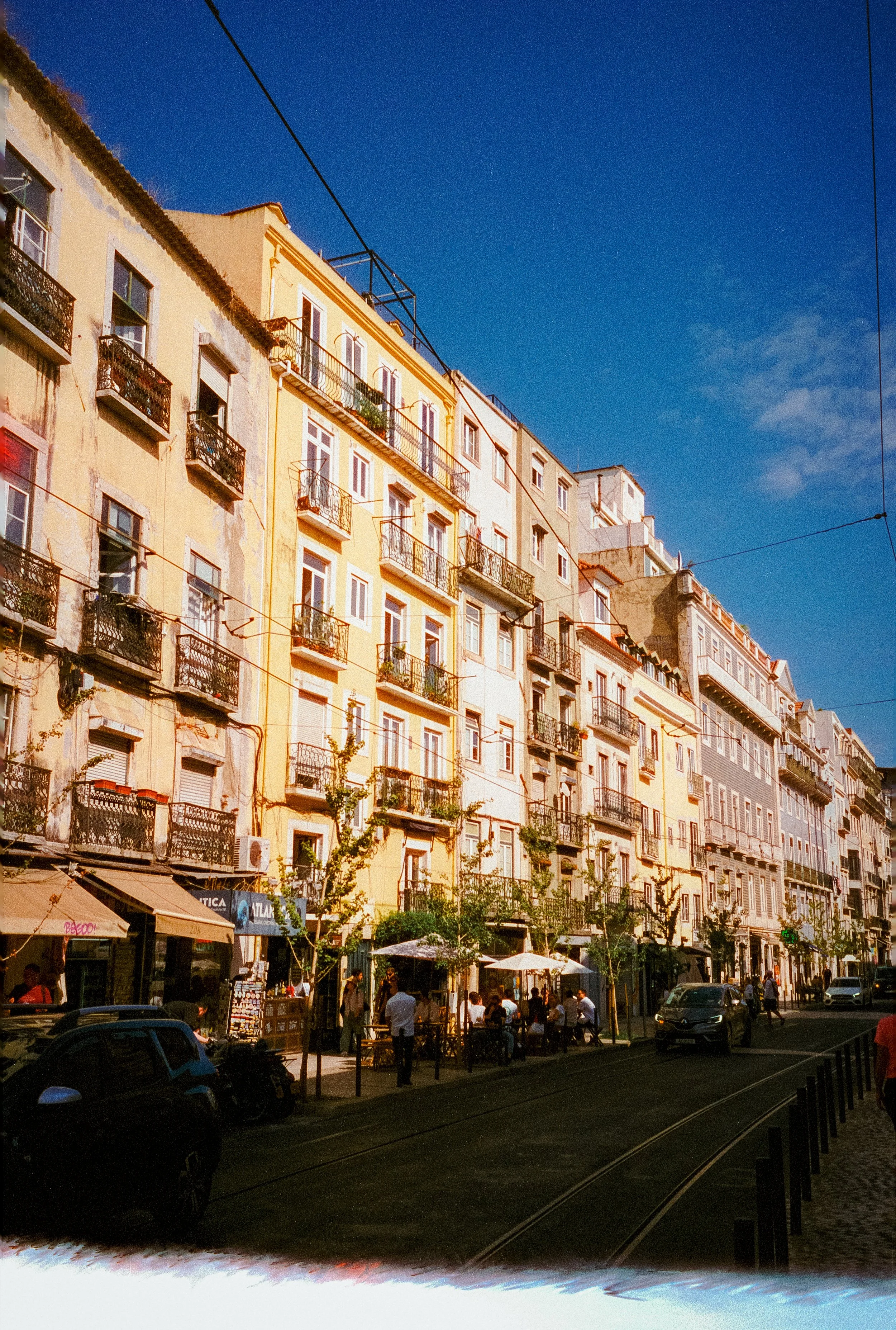 A bustling city street scene with multi-story apartment buildings, outdoor cafes, people walking, and cars parked along the street under a clear blue sky.