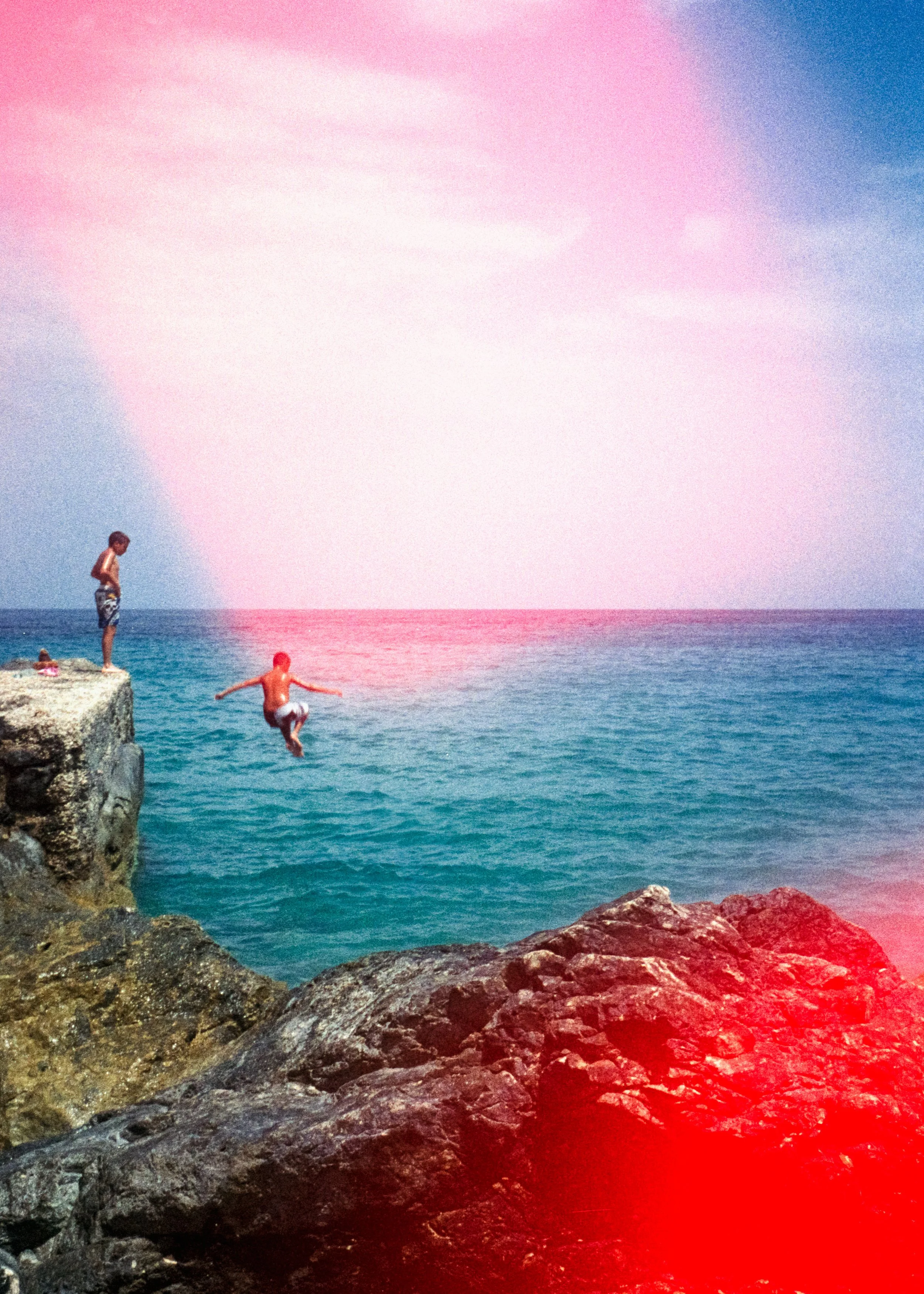 A boy jumping off a rocky ledge into the ocean at sunset, with a cloud formation resembling a rainbow in the sky.