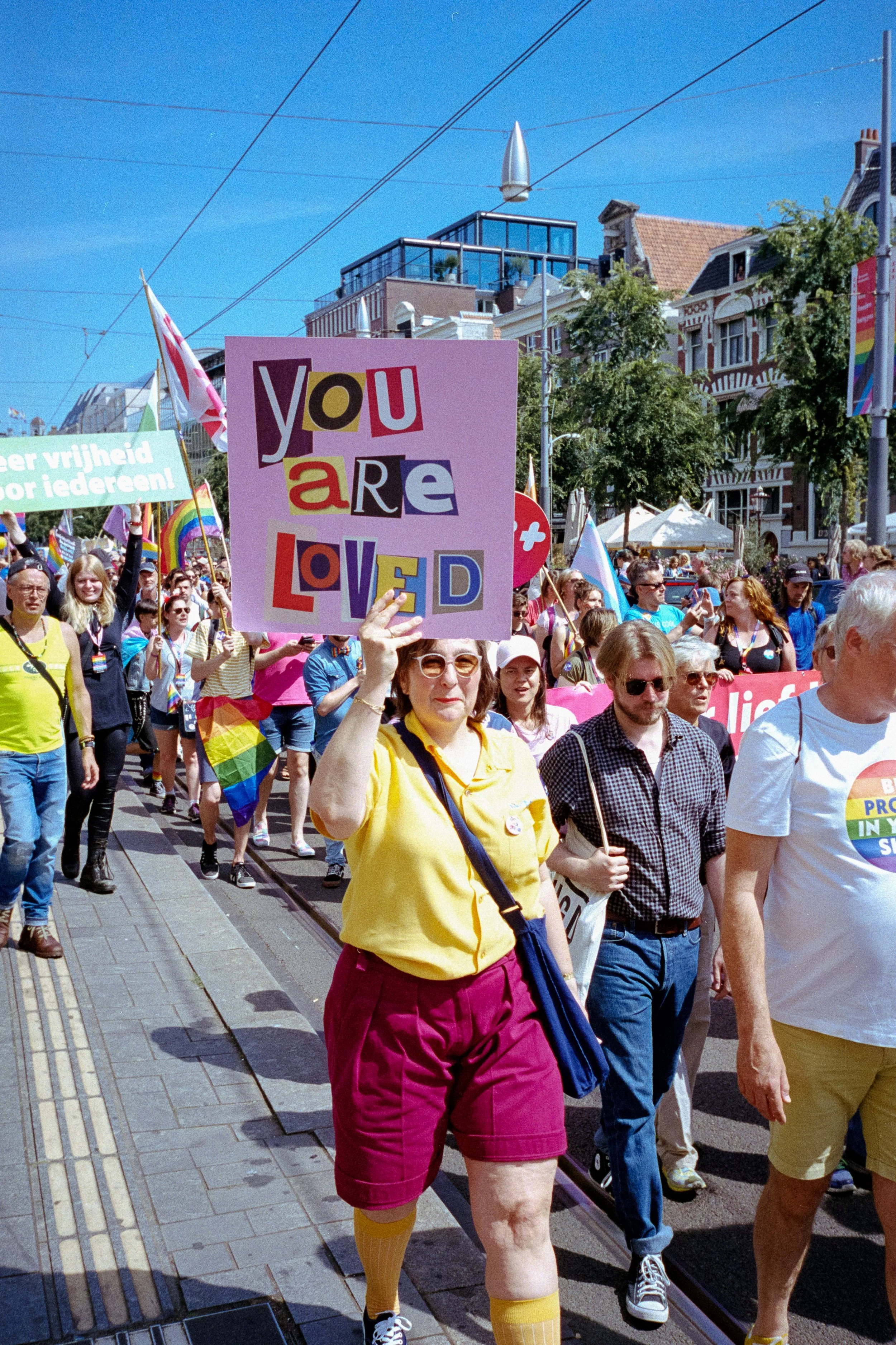 A pride parade with people walking on the street, holding rainbow flags and signs, including one that says "You are loved.