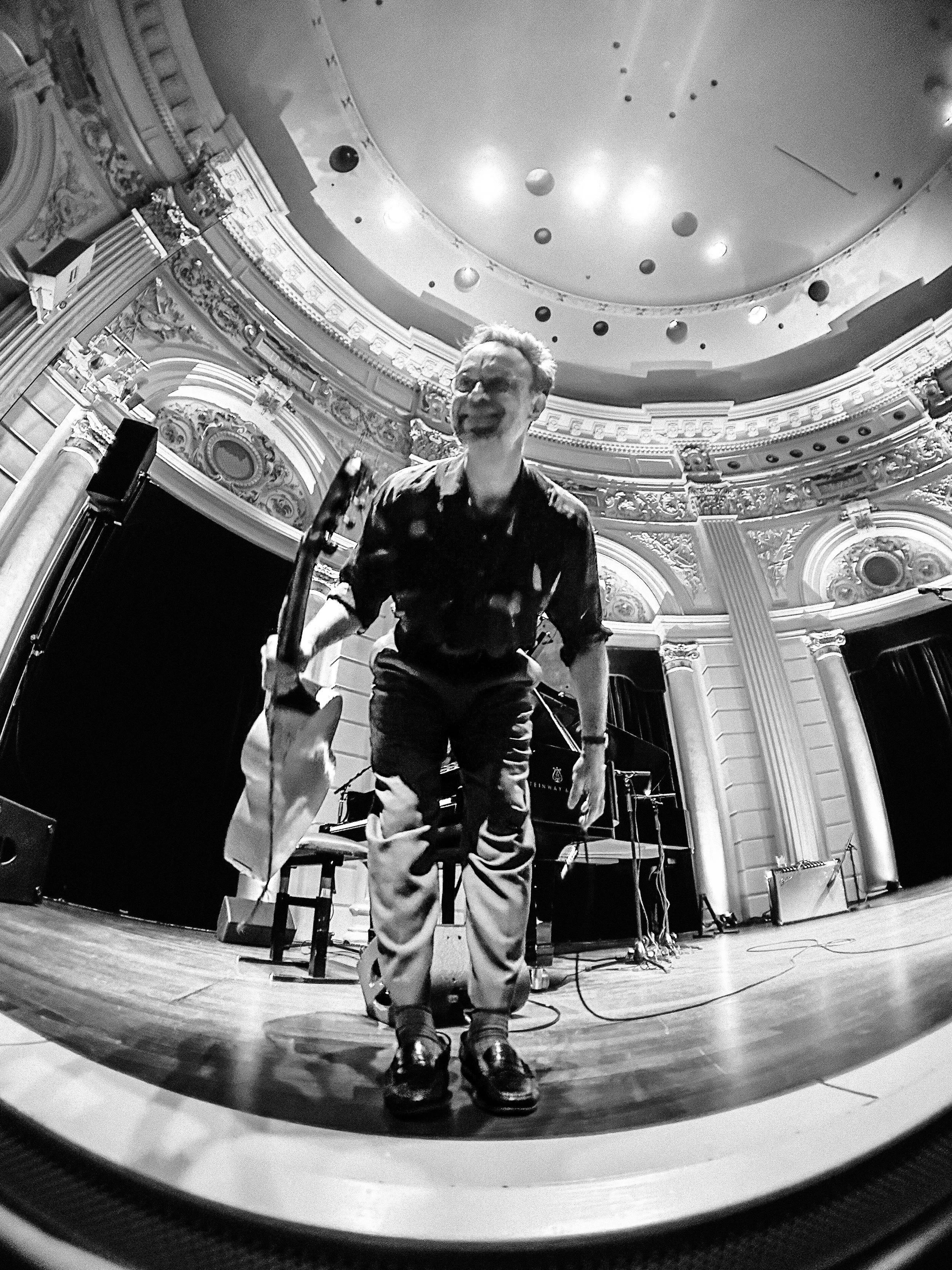 A man standing on a stage in a grand, ornate theater with intricate decorations, holding a percussion instrument, possibly a conga or drum, in front of a grand piano, with stage equipment and microphones nearby.