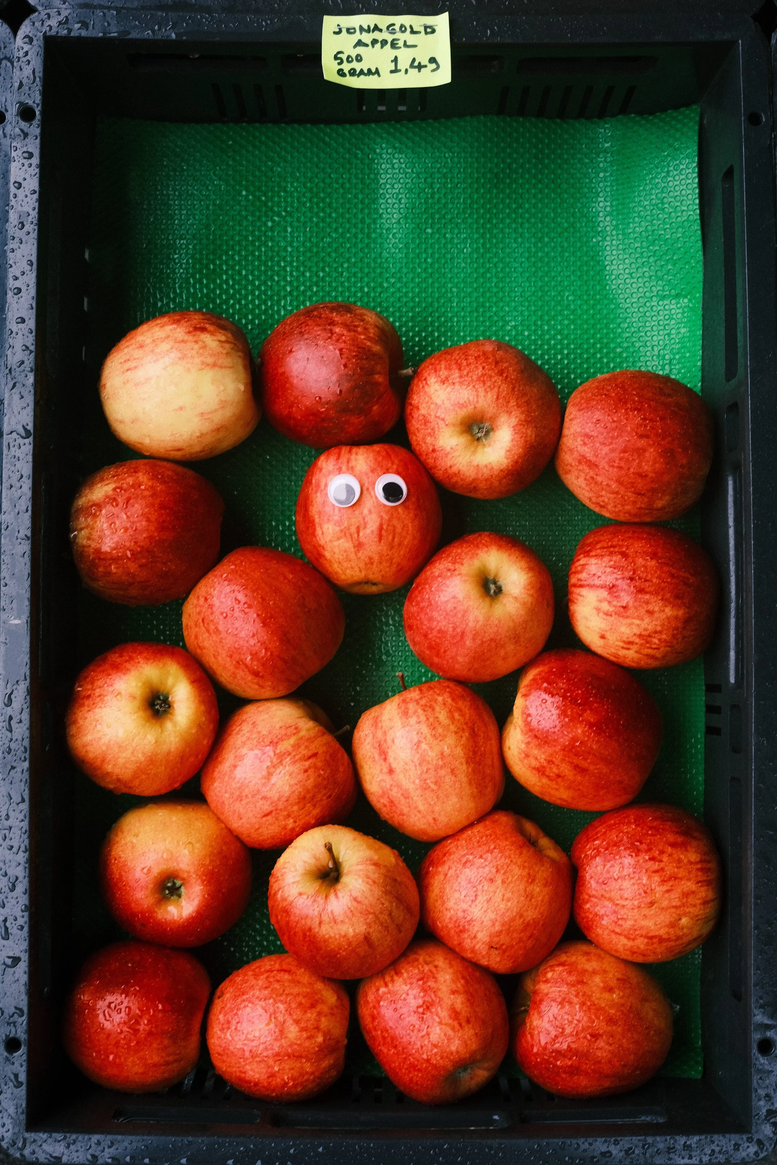 A black plastic crate filled with red and yellow apples on a green textured mat, with a yellow sticky note on top indicating they are 'Jonagold apples' weighing 500 grams each, priced at 1.49.