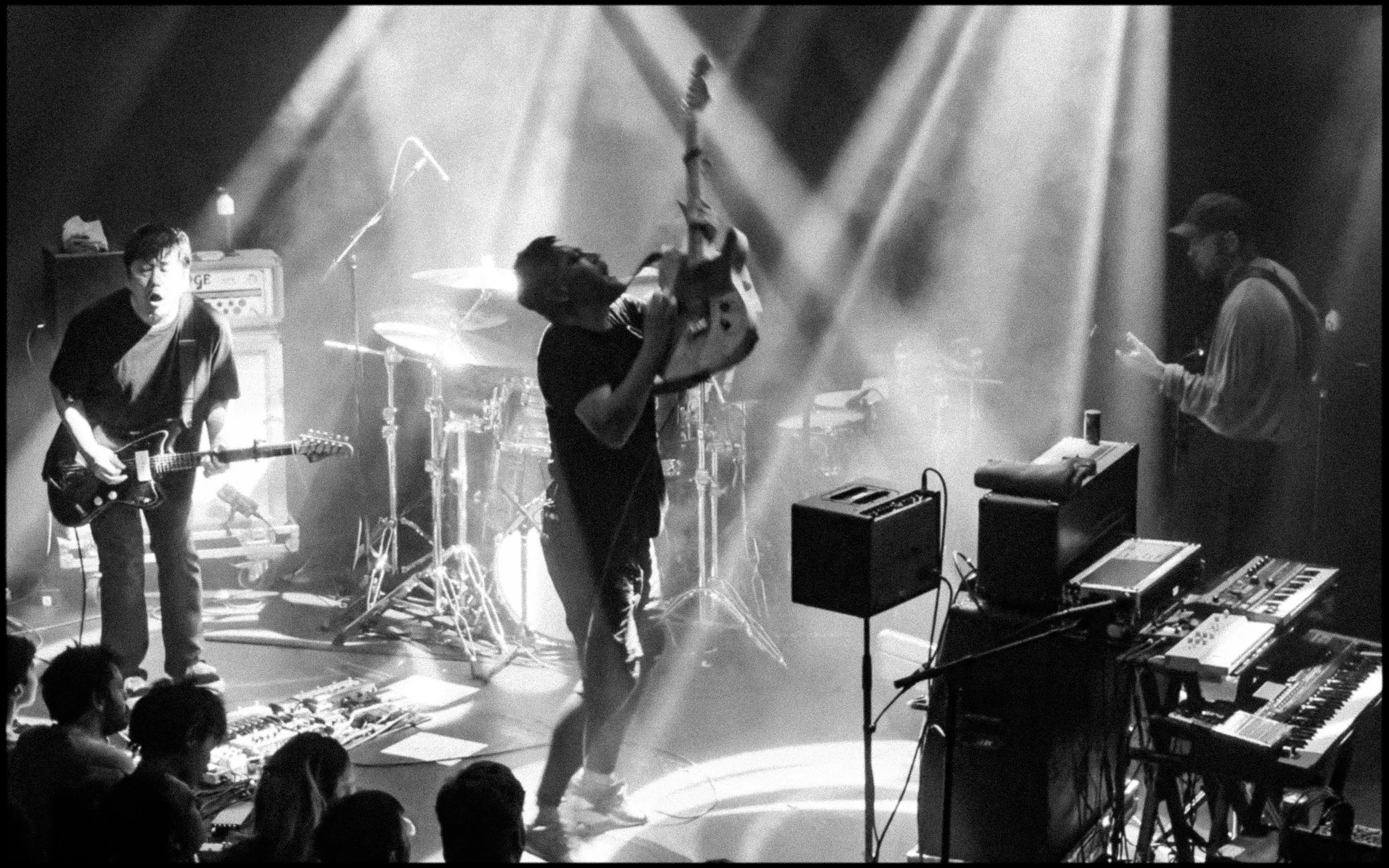 Black and white photo of a live music performance with three musicians on stage, guitars and electronic equipment, audience members in the foreground.