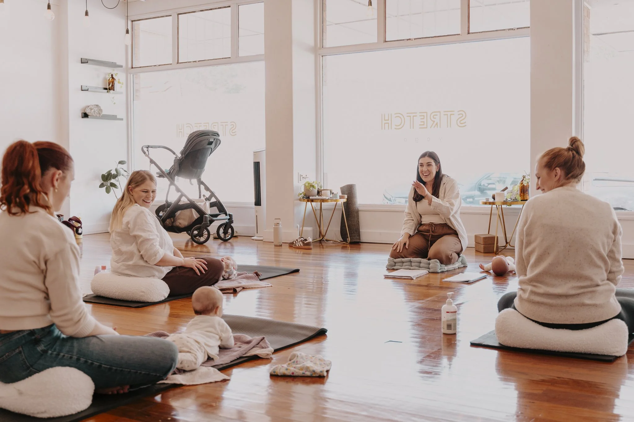 Women and babies participating in a prenatal yoga class in a bright, spacious studio with large windows, wooden floors, and minimal decor.