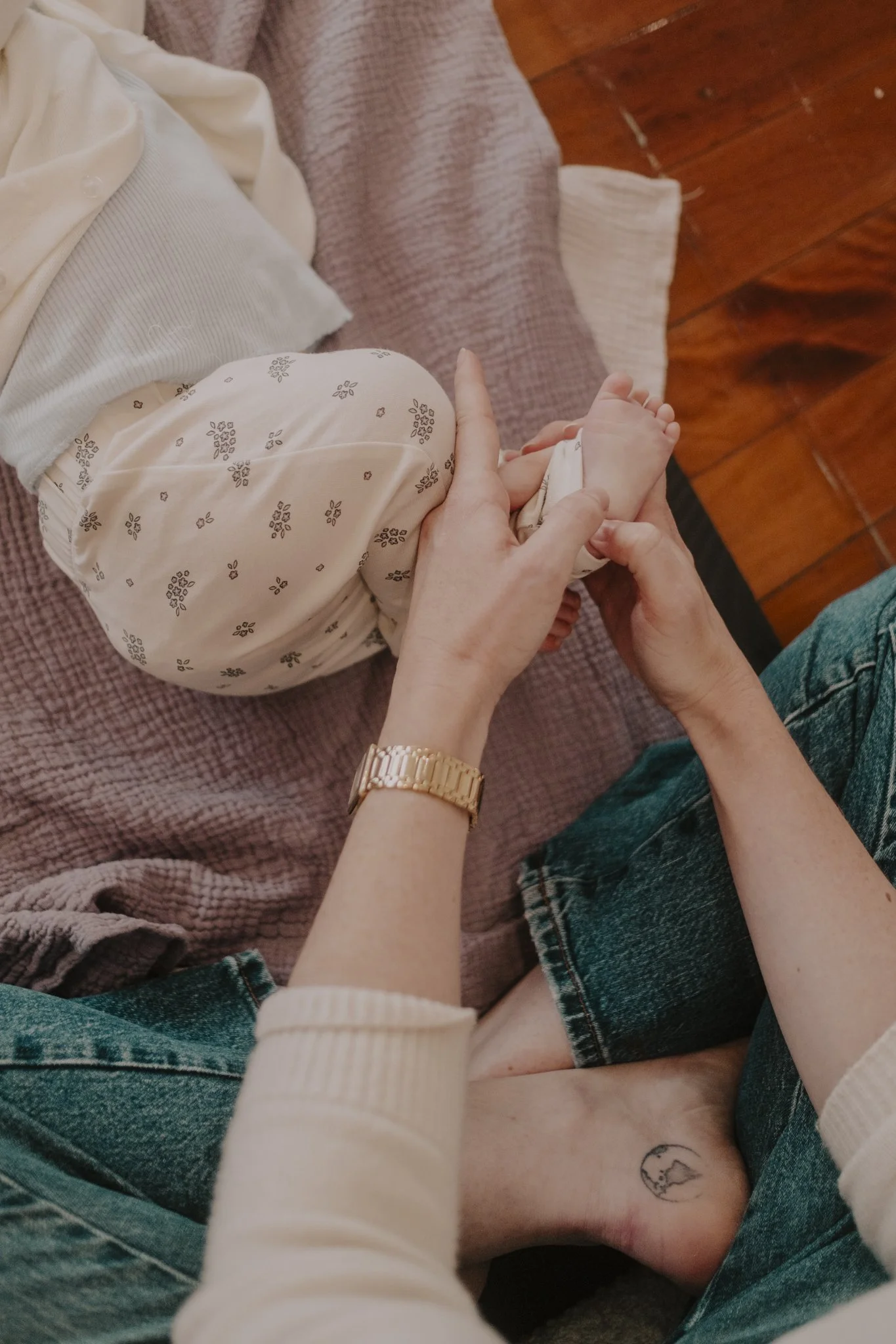A person holding a baby's foot, with a tattoo of a moon on their ankle. The baby is lying on a bed with a pink blanket, and the person is wearing a gold wristwatch and blue jeans.