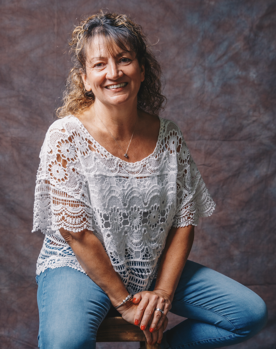 A woman with curly light brown hair wearing a white lace crochet top and light blue jeans, sitting on a stool against a textured brown background, smiling at the camera.